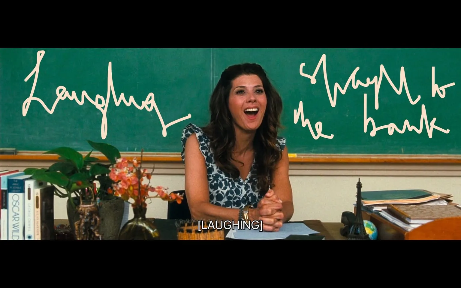 The image depicts a classroom setting with a woman seated at a desk, appearing to be engaged in a moment of laughter. She has long, wavy hair and is wearing a sleeveless dress with a blue and white floral pattern. Her expression is joyful, with her mouth open in a smile, and her hands are clasped together on the desk in front of her.

In the background, there is a green chalkboard, which is partially visible. The chalkboard features handwritten text that appears to be in a casual, flowing script. The words "Laughing" and "ShwayM to the bank" are prominently displayed, suggesting a playful or humorous context. The text is written in a light color, contrasting with the dark green of the chalkboard.

On the desk, there are various items visible, including a small potted plant with pink flowers, a stack of books, and a decorative item that resembles a globe. There is also a small Eiffel Tower figurine, adding a touch of decor to the workspace. The overall environment suggests a casual and friendly classroom atmosphere, likely during a light-hearted moment in a lesson or discussion.

The lighting in the image appears bright, contributing to the cheerful mood of the scene. The classroom is organized, with the desk neatly arranged, indicating a professional yet relaxed educational setting.