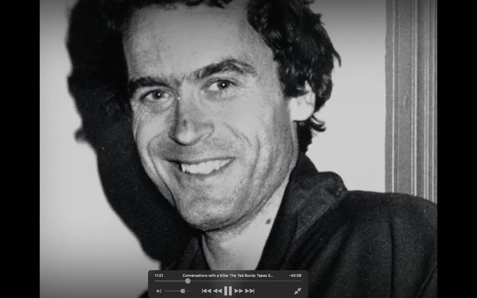 The image features a black-and-white photograph of a man with dark, slightly curly hair and a light complexion. He is smiling broadly, displaying a set of straight teeth. His facial expression appears friendly and approachable. The man has prominent eyebrows and a slight stubble on his face. 

He is wearing a dark-colored jacket, which is partially visible in the image. The collar of the jacket is turned down, revealing a light-colored shirt underneath. The background is plain and light, possibly white, which contrasts with his darker clothing. There are no visible logos, text, or other identifiable features in the background.

The lighting in the image creates a soft shadow on the left side of his face, enhancing the contours of his features. The photograph appears to be a close-up, focusing primarily on his face and upper torso. The overall composition is centered on the man, with no other objects or people present in the frame. 

The image does not contain any readable text, headlines, or signs, and there are no visible brands or products. The setting does not provide specific context, as it appears to be a simple portrait without any identifiable location or event indicators.