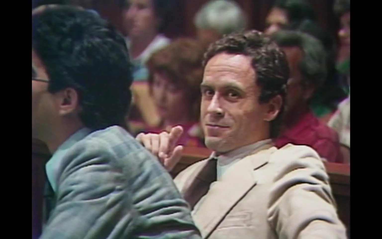 The image depicts a courtroom scene featuring two men seated at a table. The man on the left has dark, curly hair and is wearing a gray suit with a light blue shirt. He appears to be looking down or slightly away from the camera. The man on the right has light brown, wavy hair and is dressed in a light-colored suit, possibly beige, with a white shirt and a dark tie. He is leaning back in his chair, looking directly at the camera with a slight smile and a raised finger, suggesting a gesture of acknowledgment or confidence.

In the background, there are several individuals visible, including a mix of men and women. The audience appears to be engaged, with some looking towards the front of the courtroom. The setting is characterized by wooden paneling and a formal atmosphere typical of a courtroom, with rows of seats filled with spectators.

The lighting in the image is somewhat dim, contributing to the serious tone of the environment. The overall composition captures a moment during a legal proceeding, with a focus on the two men at the forefront, particularly the one with the light suit who is making eye contact with the camera. There are no visible text, signs, or logos in the image. The scene conveys a sense of tension and formality, typical of judicial settings.