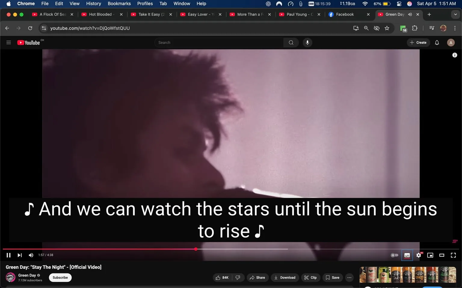 The image shows a scene from a YouTube video titled "Stay The Night" by Green Day, indicated by the logo and channel name at the bottom. The video is playing, and the timestamp shows it is at 1 minute and 57 seconds out of a total of 4 minutes and 38 seconds. 

In the foreground, there is a close-up of a person, likely a musician, who appears to be singing or performing. The individual has short, tousled hair and is partially turned to the side, with their mouth near a microphone. The background is blurred, featuring soft, muted colors, predominantly shades of pink and purple, which create a dreamy atmosphere.

At the bottom of the image, there are subtitles displayed in white text against a black background, reading: "And we can watch the stars until the sun begins to rise." The text is large and clear, making it easy to read.

The YouTube interface is visible at the top, showing various tabs and bookmarks, indicating that the user is on a computer. The video controls, including options to like, share, download, and clip, are also visible beneath the video. The screen shows a collection of beverage cans at the bottom, suggesting a casual viewing environment.

Overall, the image captures a moment from a music video, focusing on the performance aspect while providing context through the YouTube interface.