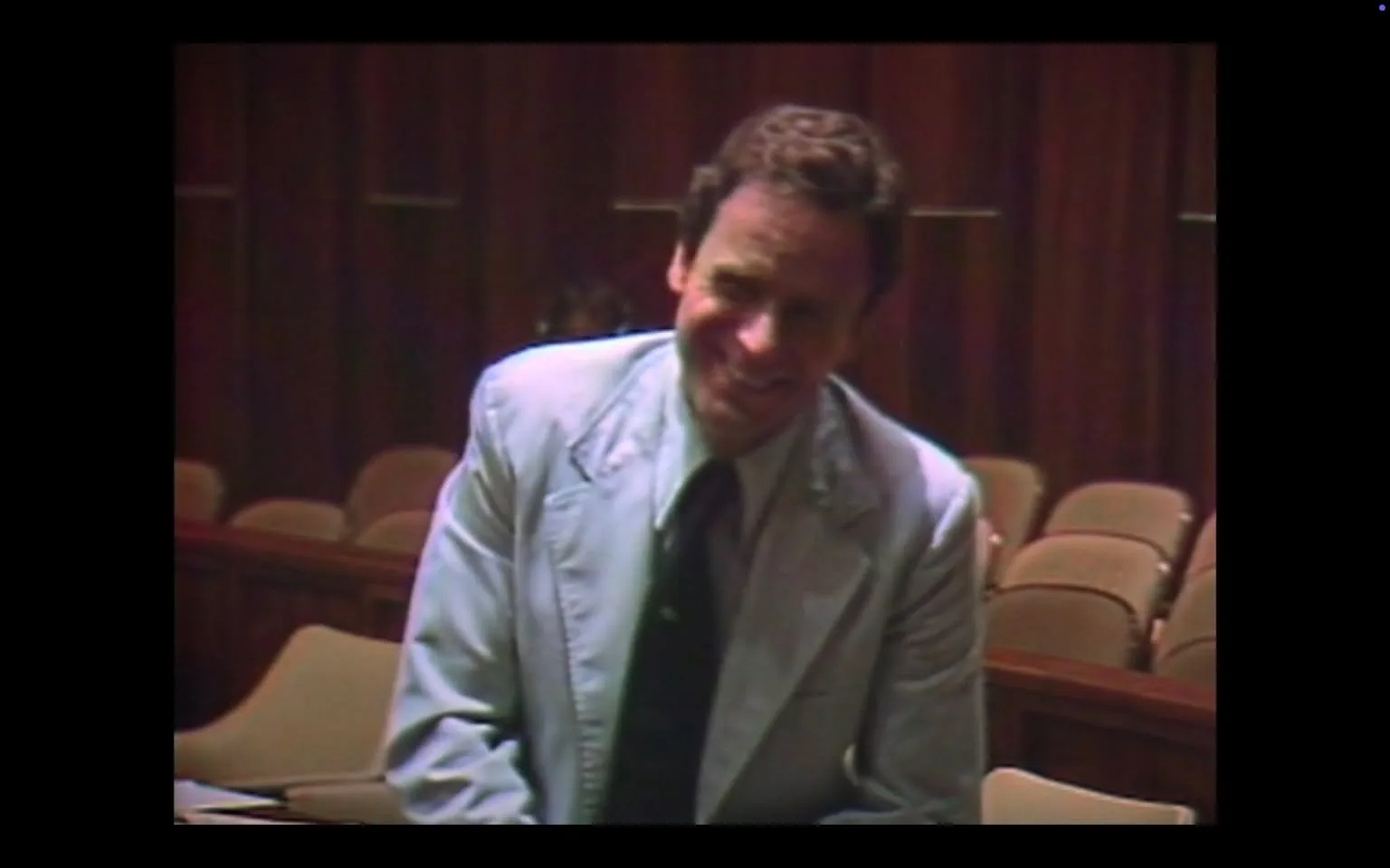 The image depicts a man in a light blue suit, standing in what appears to be a courtroom or a similar formal setting. He has a broad smile on his face, suggesting a light-hearted or jovial demeanor. His hair is dark and slightly wavy, and he appears to be in his 30s or 40s. The suit jacket has a notch lapel and is paired with a darker tie, which contrasts with the light color of the suit. 

The background features wooden paneling, which gives the environment a warm, traditional feel. There are rows of beige chairs visible behind him, indicating that this space is designed for an audience or jury. The lighting is somewhat dim, contributing to a serious atmosphere typical of a courtroom setting. 

No readable text, signs, or logos are present in the image. The man is positioned slightly to the left of the frame, and his body language suggests he is engaged in conversation or reacting to something humorous. The overall composition focuses on him, with the background providing context but not drawing attention away from his expression. 

The image captures a moment that seems to blend a formal environment with a personal interaction, highlighting the contrast between the serious setting and the man's cheerful demeanor.