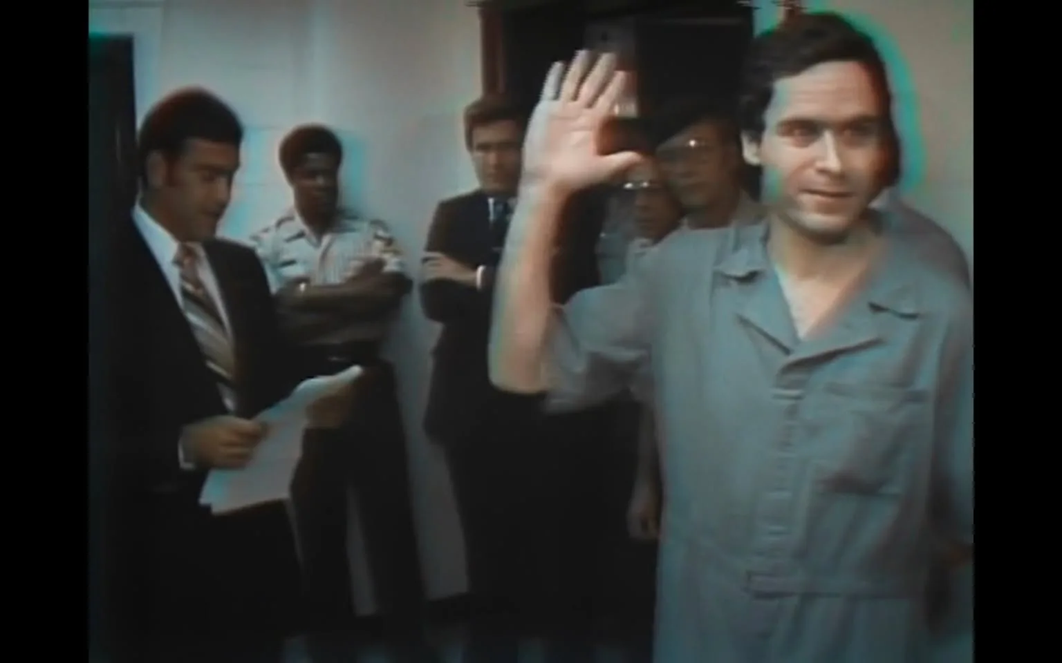 The image depicts a group of individuals in a room, likely during a press conference or a similar event. The focal point is a man in the foreground, who is raising his right hand in a gesture that could be interpreted as a wave or a sign of acknowledgment. He is wearing a light gray jumpsuit with a collar and a pocket on the left side. His hair is dark and slightly tousled, and he has a neutral expression on his face.

In the background, there are several other men standing in a semi-circle. One man, positioned to the left of the focal individual, is dressed in a dark suit with a white shirt and a patterned tie, holding a piece of paper in his left hand. He appears to be reading or preparing to speak. Another man, wearing a striped shirt, stands with his arms crossed, looking towards the focal individual. The remaining individuals in the background are dressed in various styles, including a man in glasses and a dark suit, and another in a light-colored shirt.

The setting appears to be indoors, with white walls and a doorway visible in the background. The lighting is somewhat dim, contributing to a serious atmosphere. There are no visible logos, signs, or text in the image. The overall composition suggests a formal or significant moment, likely related to a legal or newsworthy event.