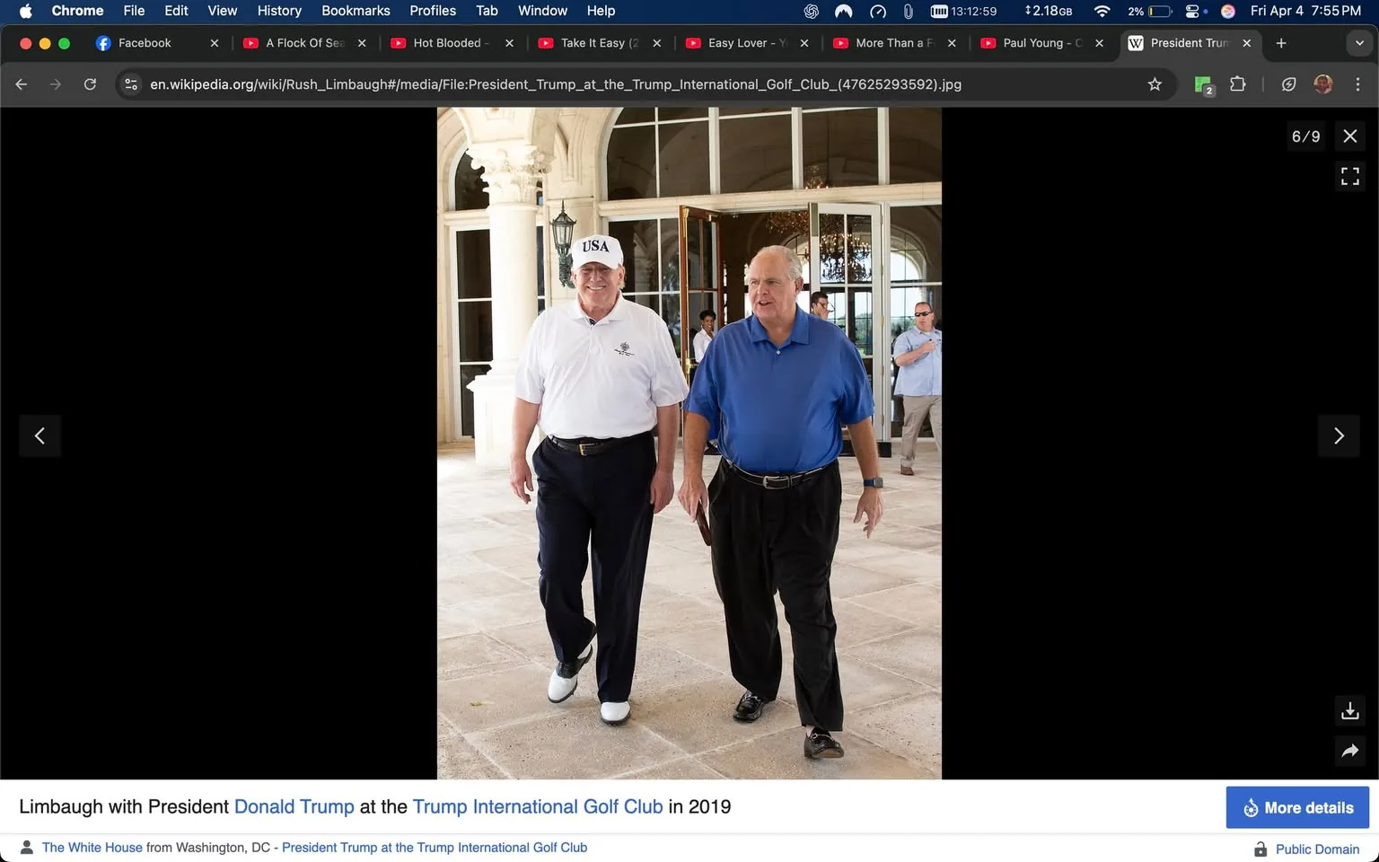 The image depicts two men walking side by side in a well-lit, spacious area, likely part of a golf club or resort. The background features large glass doors and windows, allowing natural light to flood the space. The flooring appears to be made of light-colored tiles, and there are decorative columns visible, suggesting an elegant architectural style.

On the left, one man is wearing a white polo shirt with a logo on the left side, dark pants, and white golf shoes. He is also wearing a white cap with "USA" printed on the front. His expression is neutral, and he is looking slightly to the right as he walks.

The man on the right is dressed in a blue polo shirt and black pants, with black shoes. He has a relaxed posture and is looking directly ahead. Both men are walking casually, with their arms relaxed at their sides.

In the background, there are additional individuals visible, including a man in a light blue shirt standing slightly behind the two men in the foreground. The setting appears to be a social or recreational environment, likely related to golf or leisure activities.

No specific logos, signs, or text are prominently visible in the image aside from the cap worn by the man on the left. The overall atmosphere of the scene suggests a friendly and informal outing.