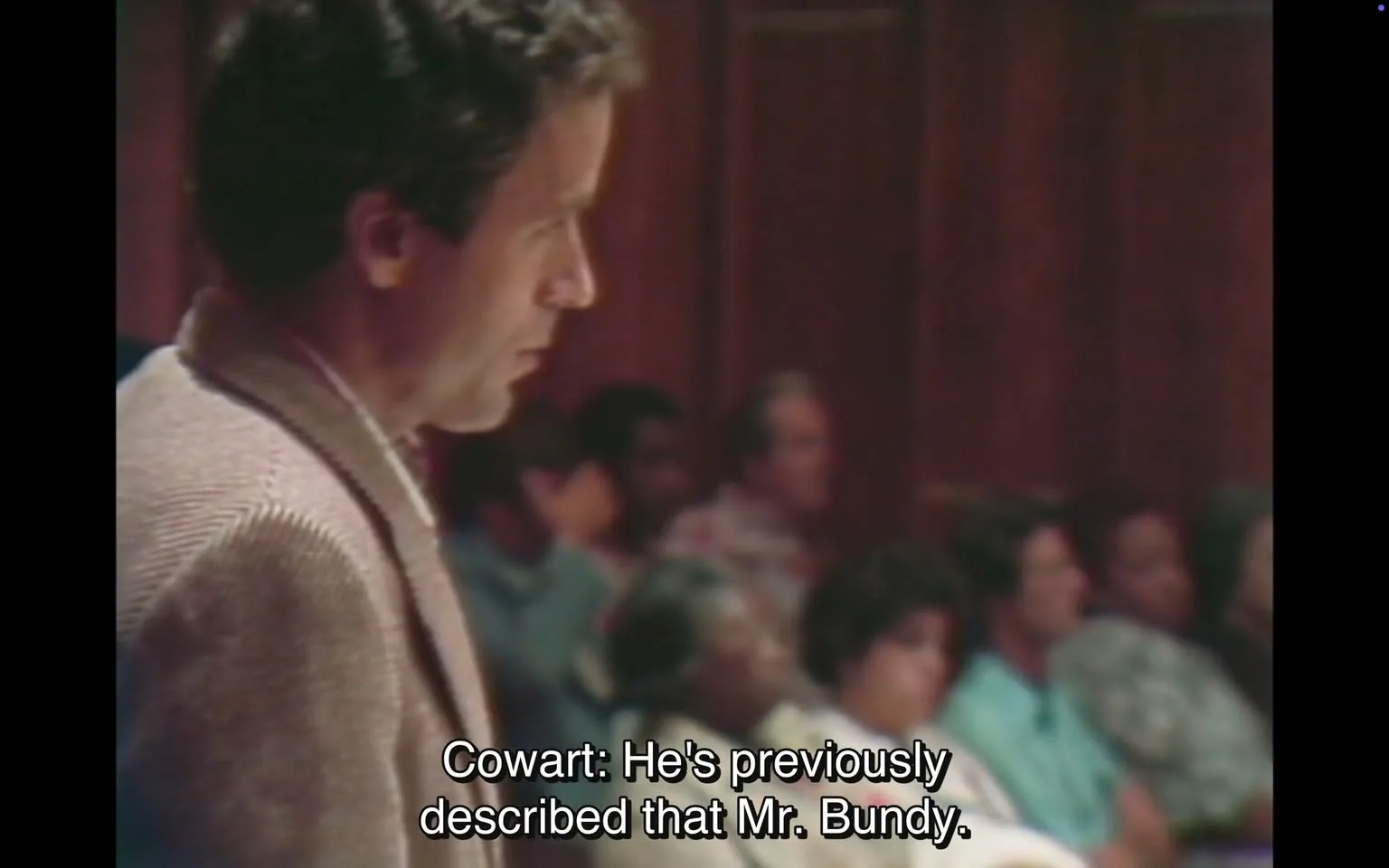 The image depicts a courtroom scene. On the left side, a man is standing, appearing to be in mid-sentence, possibly addressing the court. He has dark, slightly curly hair and is wearing a light-colored suit jacket with a textured pattern. His expression is serious, and he is looking slightly downward, suggesting he is focused on his statement or the proceedings.

In the background, a group of people is seated, likely members of the audience or jury. They are partially obscured, but several individuals can be seen. Some are wearing casual clothing, including a blue shirt and a patterned shirt. The audience appears to be engaged, with some individuals looking towards the speaker.

The courtroom has wooden paneling, giving it a formal and traditional appearance. The lighting is somewhat dim, contributing to the serious atmosphere of the setting. The text at the bottom of the image reads: "Cowart: He's previously described that Mr. Bundy," indicating that the man speaking is likely referencing a previous description related to a person named Bundy.

Overall, the scene conveys a formal legal environment, with a focus on the speaker and the attentive audience. The details of clothing and expressions suggest a serious discussion is taking place, likely related to a legal matter.