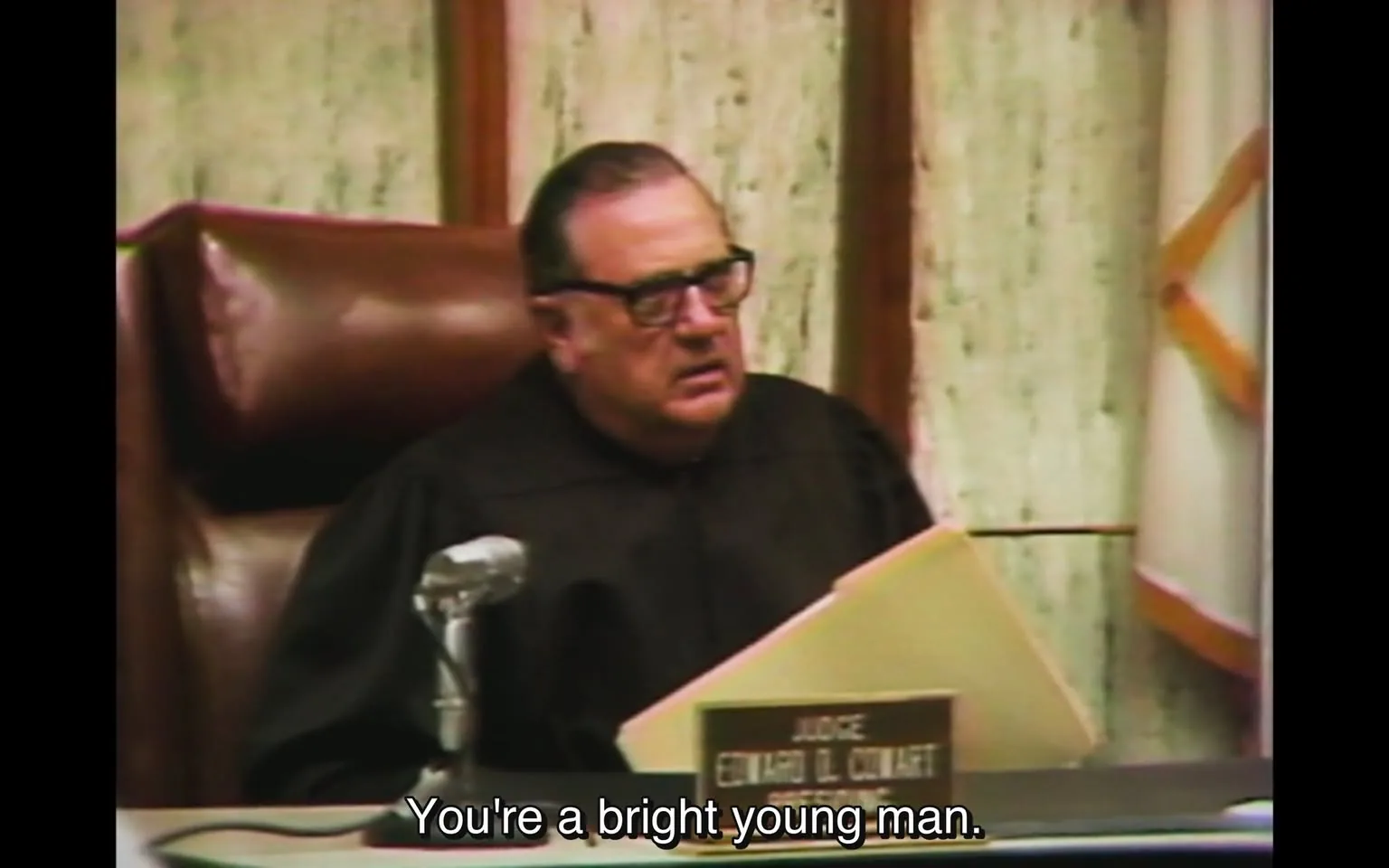 The image depicts a courtroom setting with a judge seated at the bench. The judge is a middle-aged man with glasses, wearing a black robe. He has a serious expression on his face, indicating he is engaged in a discussion or deliberation. His hair is dark and slicked back, and he appears to be looking intently at a file or document in his hands.

In front of the judge, there is a microphone on the bench, suggesting that the proceedings are being recorded or broadcast. The judge's nameplate is visible, reading "JUDGE EDWARD D. CULHANE," indicating his identity and position. The background features a textured wall that appears to be made of a light-colored material, possibly marble or a similar stone, with wooden paneling framing the area. 

The judge is holding a folder, which is partially open, but the contents are not visible. The overall atmosphere of the courtroom is formal and serious, typical of judicial proceedings. The lighting is soft, contributing to the solemnity of the setting. There are no other individuals visible in the frame, focusing attention solely on the judge and his actions. 

The text at the bottom of the image reads, "You're a bright young man," suggesting a statement made by the judge, possibly addressing someone not visible in the image. The overall composition emphasizes the authority of the judge within the courtroom environment.