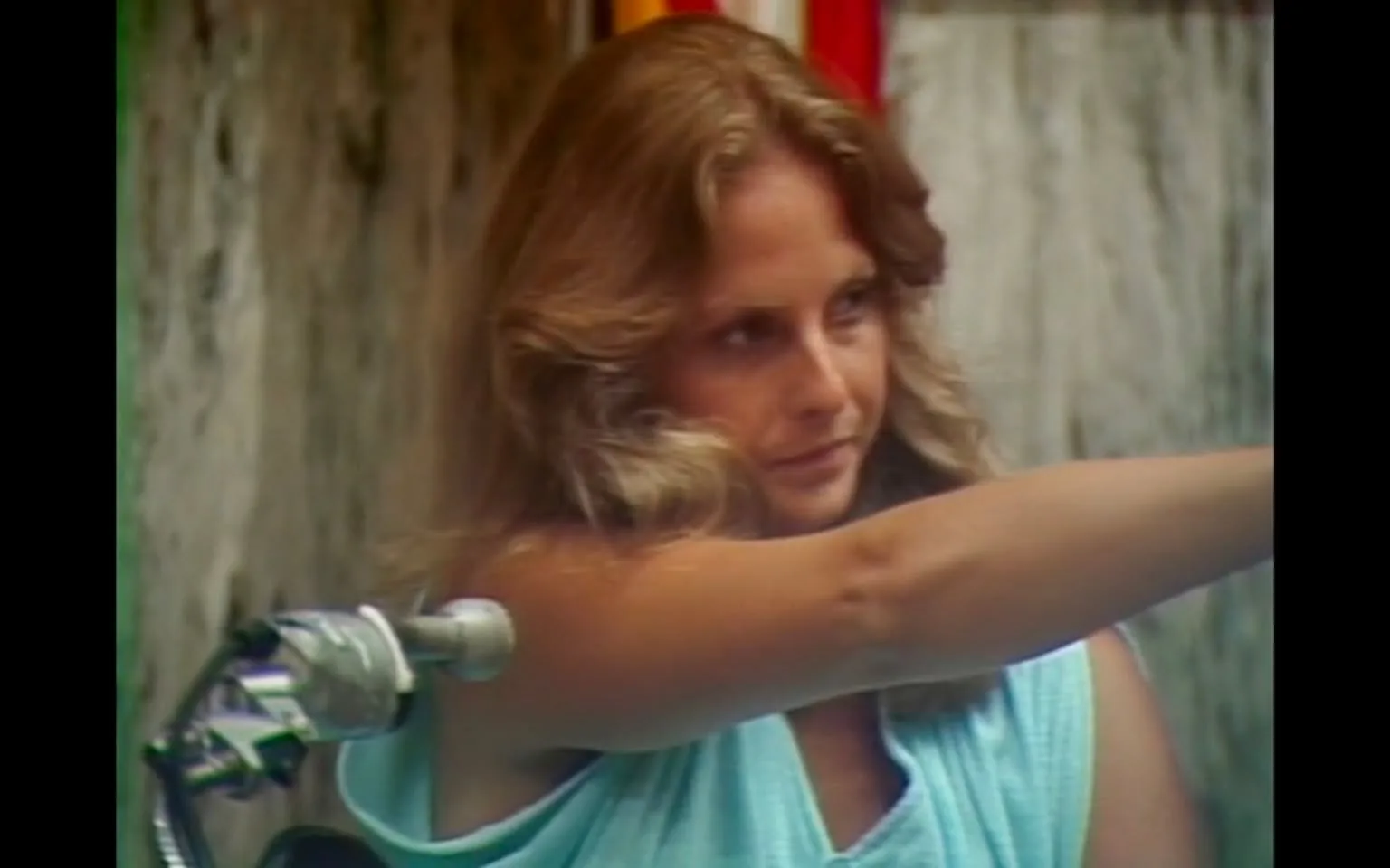 The image features a woman with long, wavy hair that appears to be light brown or blonde. She is wearing a light blue, sleeveless top that has a loose fit, exposing her arms. The woman is positioned slightly to the right of the frame, with her body angled forward as she extends her right arm outward. Her facial expression is serious and focused, with her gaze directed towards the camera or an unseen object in front of her.

In the background, there is a textured wall that appears weathered, possibly made of concrete or stone, with some discoloration and markings. The colors of the wall are muted, primarily gray with hints of brown. There are also some vertical stripes of red and yellow visible in the background, suggesting the presence of flags or banners, but the details are not clear.

To the left of the woman, there is a motorcycle or scooter partially visible, with a chrome handlebar that reflects light. The handlebar has a grip and a lever, indicating it is part of a motorized vehicle. The setting appears to be outdoors, possibly in an urban environment, but no specific location or identifiable landmarks are visible.

Overall, the image captures a moment of action or intent, with the woman seemingly engaged in a task or activity, though the exact context is not clear from the visual alone.