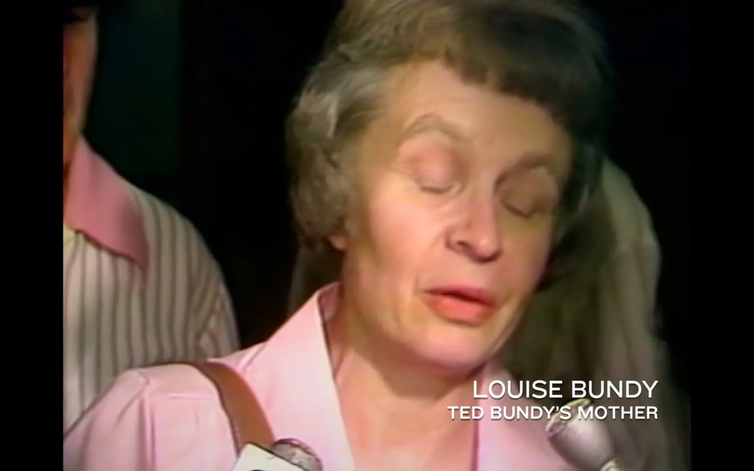 The image features a woman identified as Louise Bundy, who is described as Ted Bundy's mother. She has short, wavy hair that is grayish in color and styled in a way typical of the 1970s. Her facial expression appears serious, and her eyes are partially closed, suggesting she may be in the midst of speaking or reflecting. She is wearing a light pink blouse with a collar that has a darker pink trim, which is buttoned up. A brown shoulder bag is visible on her left shoulder.

In the background, there is a person wearing a striped shirt, which is primarily white with thin vertical stripes. The setting appears to be indoors, possibly at a press conference or a public event, given the presence of microphones and the way the woman is positioned as if addressing an audience or reporters.

The bottom right corner of the image contains text that reads "LOUISE BUNDY" and "TED BUNDY'S MOTHER," indicating her identity and relationship to Ted Bundy. There are no visible logos, trademarks, or other identifiable brands in the image. The overall tone of the image is somber, reflecting the serious context surrounding the individuals involved. 

The background is somewhat blurred, focusing attention on Louise Bundy, while the lighting appears to be soft, typical of a media setting. There are no additional objects or signs present that provide further context to the location or event.