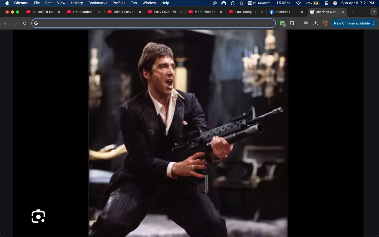 The image depicts a male figure in a dramatic pose, holding a firearm. He appears to be in a stylized, cinematic setting, likely from a film. 

**Clothing and Appearance:**
- The individual is dressed in a black pinstripe suit, which is tailored and fitted. 
- He has a white dress shirt underneath, with the collar slightly open and a hint of a white undershirt visible.
- The suit jacket has a pocket square, which is red, adding a pop of color to the outfit.
- His hair is dark and styled in a slicked-back manner, with a slightly tousled look.
- The expression on his face is intense, with his mouth open as if shouting or expressing strong emotion. His eyes are wide, conveying a sense of urgency or aggression.

**Action:**
- He is positioned in a dynamic stance, with his legs spread apart and knees slightly bent, suggesting movement or readiness.
- The firearm he is holding is a black assault rifle, with a prominent barrel and a tactical design. He grips it firmly with both hands, indicating he is prepared for action.

**Setting:**
- The background is dimly lit, with a luxurious, opulent feel. There are ornate chandeliers visible, suggesting a grand interior space, possibly a mansion or a lavish room.
- The floor appears to be carpeted, and there are hints of furniture in the background, though details are obscured by the lighting.

**Overall Context:**
- The image conveys a sense of high tension and drama, typical of action or crime genres in film. The character's pose and expression suggest a pivotal moment, possibly in a confrontation or climactic scene.

No readable text, logos, or trademarks are present in the image. The focus remains on the character and his immediate environment.