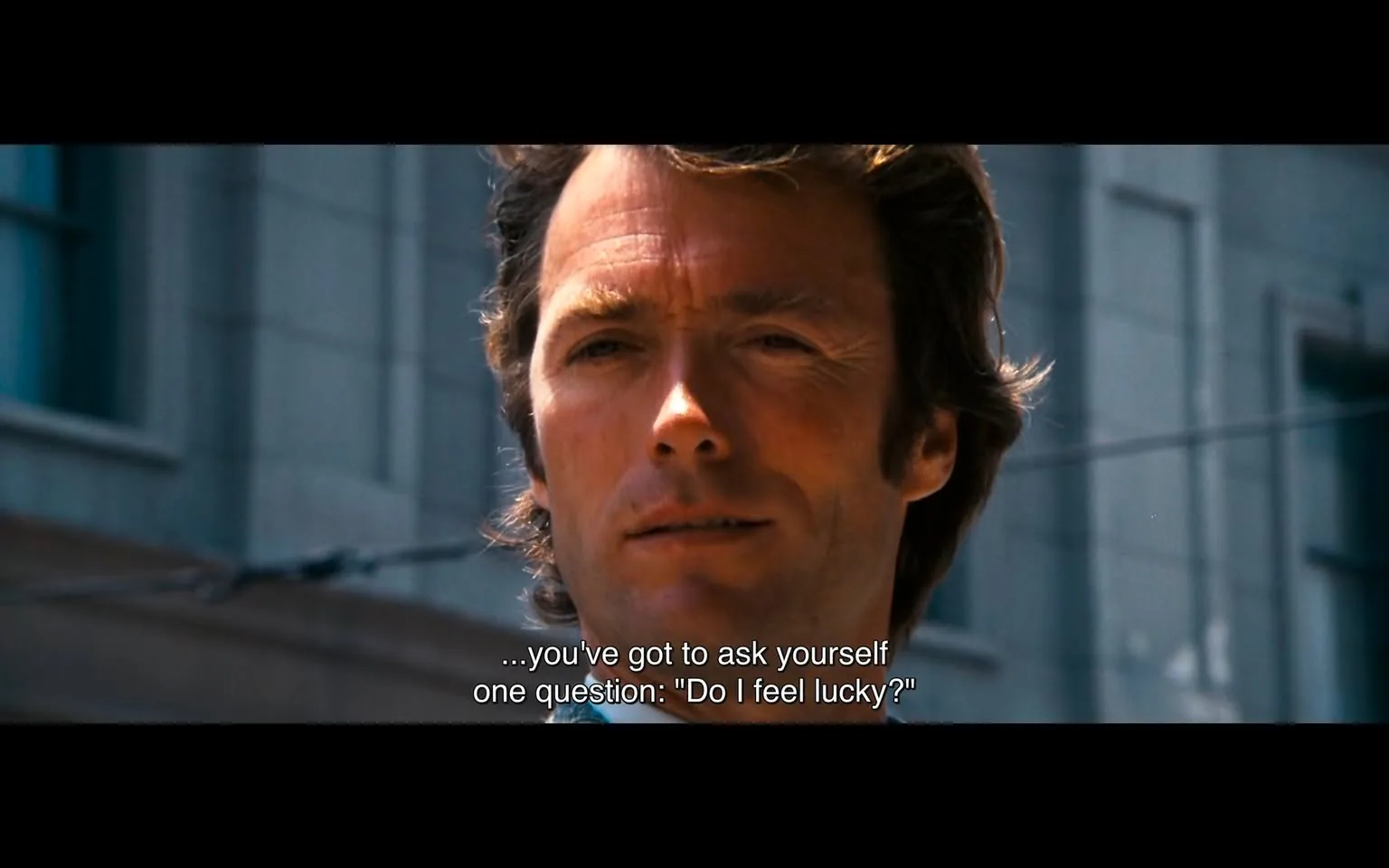 The image features a close-up of a man with light brown hair styled in a slightly tousled manner. He has a fair complexion and appears to be in his mid-30s to early 40s. His expression is serious and contemplative, with a slight furrow in his brow and a subtle smile. He is wearing a light-colored shirt, possibly blue, with a collar that is open at the neck. 

The background consists of a blurred urban setting, with buildings that have a modern architectural style. There are hints of windows and a structure that suggests a city street. The lighting appears bright, indicating that it may be daytime. 

Text is displayed at the bottom of the image, which reads: "...you've got to ask yourself one question: 'Do I feel lucky?'" This text is in a white font and is positioned centrally at the bottom of the image, contrasting against the darker tones of the man's shirt and the background.

No logos, brands, or product labels are visible in the image. The overall composition focuses on the man's face and expression, with the text adding context to the scene. The atmosphere is tense and dramatic, likely suggesting a pivotal moment in a narrative.