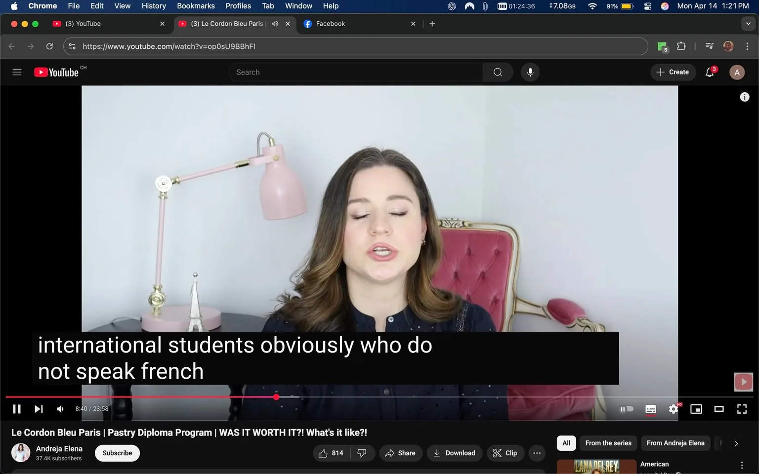 The image depicts a scene from a YouTube video featuring a woman speaking directly to the camera. She has long, wavy brown hair and is wearing a dark blue shirt with a button-up design, which has a subtle pattern of small dots. Her facial expression appears focused as she talks, and her mouth is slightly open, indicating she is in the midst of speaking.

In the background, there is a light-colored wall, and to her right, there is a pink lamp with a flexible arm, which is positioned on a desk or table. The lamp has a round base and a shade that appears to be made of metal. The desk also features a small decorative object resembling a star, which is likely a decorative piece. 

The woman is seated on a plush, pink chair with a tufted design, which adds a touch of elegance to the setting. The overall color scheme of the background is light and soft, contributing to a calm atmosphere.

At the bottom of the image, there is a black bar displaying video controls, including a play button, a timeline showing the video duration (8:40 out of 23:58), and options to share, download, or clip the video. The title of the video is visible: "Le Cordon Bleu Paris | Pastry Diploma Program | WAS IT WORTH IT?! What's it like?!" The channel name "Andrej Elena" is also displayed, along with a subscriber count of 37.4K.

Additionally, there are subtitles visible at the bottom of the image, stating: "international students obviously who do not speak french." The text is in white against a black background, ensuring it is legible. 

Overall, the image captures a moment from a personal and informative video, likely aimed at sharing experiences related to studying at Le Cordon Bleu Paris.