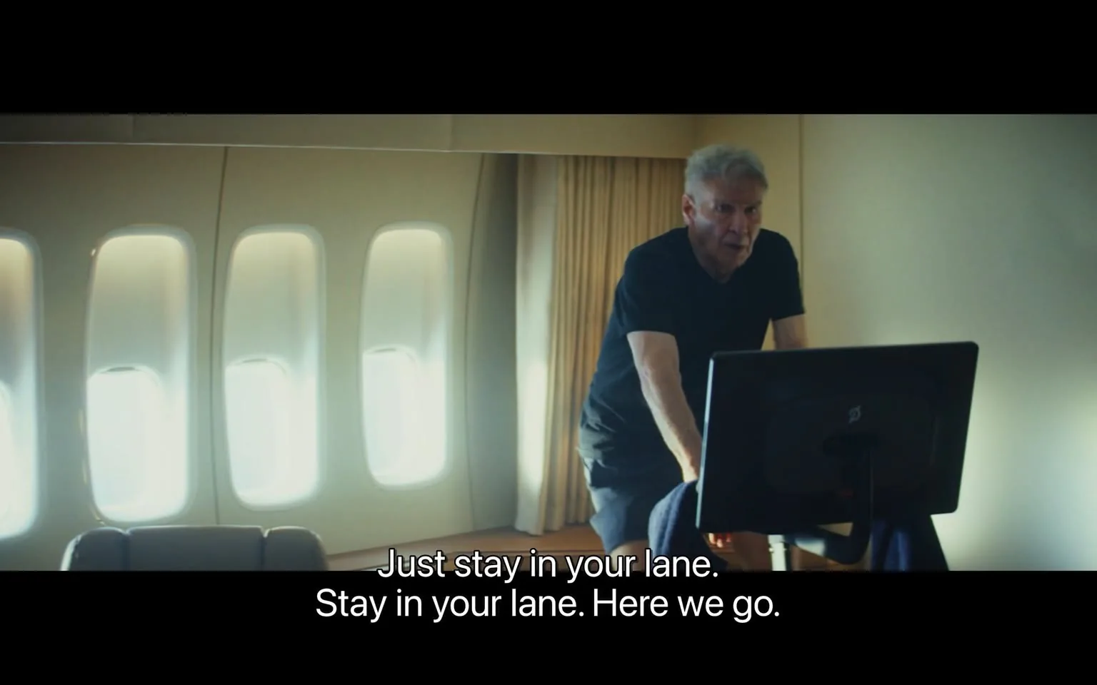 The image depicts a scene inside an aircraft, likely a private jet, characterized by a modern and spacious interior. The background features large windows with a soft, diffused light coming through, illuminating the space. There are two windows visible, both with a rounded shape, and they are positioned on the left side of the image. The walls are a light color, contributing to a bright atmosphere.

In the foreground, a man is engaged in an activity on a stationary exercise bike, which is positioned in front of a screen or monitor. The man is wearing a black short-sleeve shirt and dark pants. He has gray hair and appears to be in his late 70s or early 80s. His expression is focused, and he is leaning slightly forward, suggesting engagement in the task at hand. His right arm is raised, possibly gesturing or adjusting something on the bike or screen.

The screen in front of him is dark and appears to be a digital display, but no specific logos or text are visible on it. The exercise bike itself is modern, with a sleek design, and features a console that includes buttons or a touchscreen interface.

The text displayed at the bottom of the image reads: "Just stay in your lane. Stay in your lane. Here we go." This text is in white font against a black background, indicating it may be a subtitle or caption related to the scene.

Overall, the setting conveys a sense of activity and focus, with the man engaged in physical exercise within the confines of a luxurious aircraft environment.