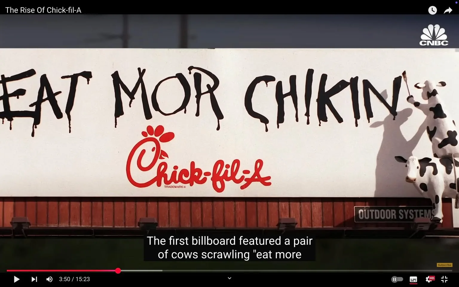 The image features a billboard with a white background and a wooden base. The main text on the billboard reads "EAT MOR CHIKIN" in large, black, dripping letters. The font appears to be hand-painted, giving it a rustic and informal look. Below this text, there is a red logo of Chick-fil-A, which includes a stylized chicken head and the brand name "Chick-fil-A" in a bold, red font. The logo is recognizable and prominently displayed.

To the right of the text, there are two cartoonish cows, one standing and one partially leaning against the billboard. The cows are painted in a classic black-and-white pattern, resembling Holstein cows. The standing cow is depicted as holding a paintbrush, suggesting it is involved in the act of painting the message on the billboard. The cows appear to be anthropomorphized, adding a humorous touch to the advertisement.

The setting appears to be outdoors, likely along a roadside or in a rural area, as indicated by the visible wooden structure of the billboard. The background is somewhat blurred, but it suggests an open space, possibly with grass or fields.

Overall, the billboard is designed to attract attention with its playful imagery and bold text, promoting Chick-fil-A in a unique and memorable way.