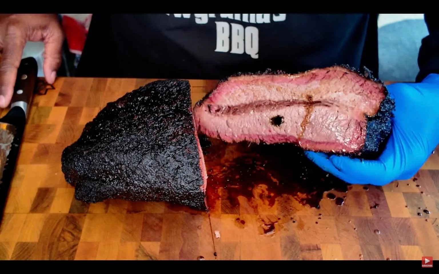 The image shows a close-up view of a large piece of brisket that has been sliced in half, revealing its interior. The brisket has a dark, charred crust on the outside, indicating it has been smoked or cooked at high temperatures. The interior of the brisket is a pinkish color, showcasing a tender texture with visible marbling of fat, which is typical for well-cooked brisket. There is a small hole in the meat, possibly from a probe used during cooking.

The brisket is placed on a wooden cutting board, which has a checkerboard pattern, indicating it is made of multiple pieces of wood. The cutting board appears to be well-used, with some visible stains and marks from previous cuts. 

In the background, a person’s hand is visible, wearing a blue glove, which is holding the sliced brisket. The hand appears to be steady, indicating that the person is likely preparing to serve or present the meat. A knife is also visible on the cutting board, suggesting that the brisket has just been cut.

The person is wearing a black shirt with the text "Grandma's BBQ" printed on it, although the details of the shirt are somewhat obscured. The setting appears to be a kitchen or a barbecue area, with various utensils and equipment likely present but not fully visible in the frame. 

Overall, the image captures a moment of food preparation, focusing on the brisket as the main subject, highlighting its texture and cooking style.