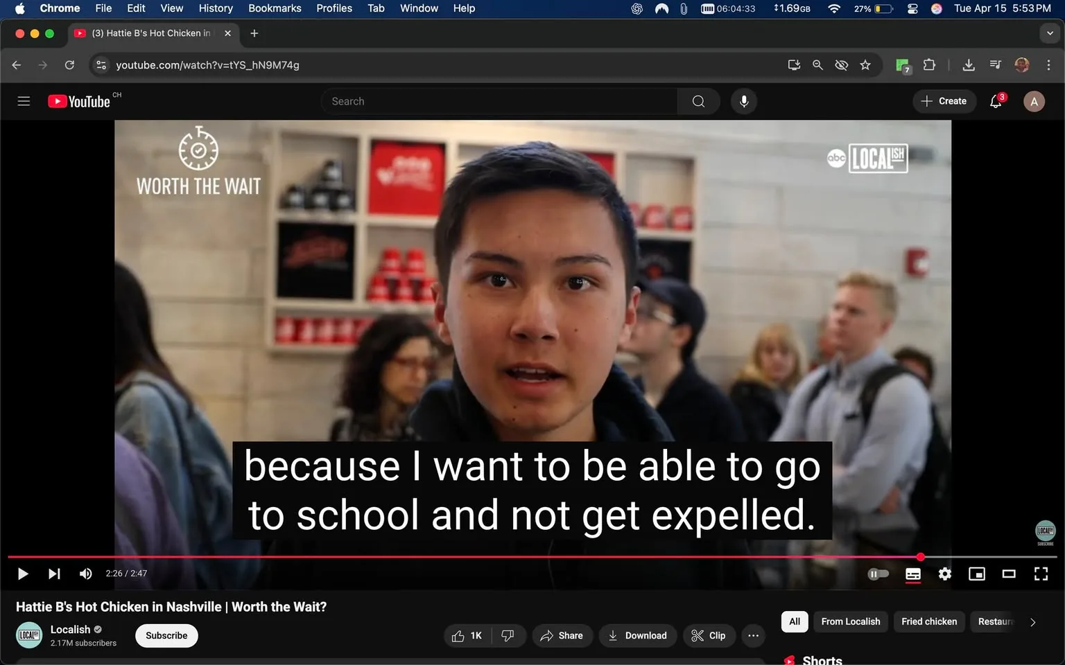 The image depicts a scene from a YouTube video titled "Hattie B's Hot Chicken in Nashville | Worth the Wait?" The video is produced by Localish and has 2.17 million subscribers. The visible portion of the video shows a young person speaking directly to the camera. 

**Text Elements:**
- The title "WORTH THE WAIT" is prominently displayed at the top of the image.
- Subtitles are visible at the bottom of the screen, reading: "because I want to be able to go to school and not get expelled."
- The YouTube interface is visible, including the video controls, with options to like, share, download, and clip the video.

**People:**
- The young person in focus has short, dark hair and is wearing a black hoodie. Their expression appears serious as they speak.
- In the background, there are several people. One person with curly hair is wearing a light blue denim jacket. Another individual, slightly blurred, has blonde hair and is wearing a dark shirt. There is also a person with a backpack, indicating a casual setting.
- The individuals in the background appear to be engaged in conversation or waiting, but their expressions are not clearly visible due to the focus on the speaker.

**Setting:**
- The setting appears to be a food establishment, likely a restaurant or food stall, given the context of the video about Hattie B's Hot Chicken, which is known for its spicy fried chicken.
- The background features a wall with shelves displaying red cups and possibly other branded items, suggesting a casual dining atmosphere.
- The overall lighting is bright, indicating an indoor environment.

**Environmental Context:**
- The scene captures a moment of interaction, likely during a busy time at the restaurant, as indicated by the presence of multiple people in the background.
- The video format suggests it is part of a series exploring local food spots, emphasizing the experience of waiting for popular food items.

This description captures the observable details without interpretation or speculation, focusing solely on the visual elements present in the image.