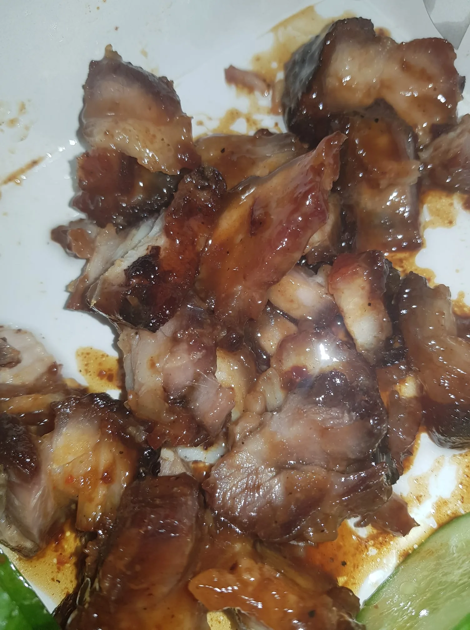 The image depicts a close-up view of a serving of cooked meat, likely barbecued or roasted. The pieces of meat are irregularly shaped and vary in size, with some showing a glossy, caramelized surface, indicating a glaze or sauce. The meat appears to be well-cooked, with a combination of dark brown and lighter shades, suggesting a mix of crispy and tender textures. 

There are visible juices pooling around the meat, which may indicate that it has been marinated or cooked with a flavorful sauce. The background is a plain white container, possibly a takeout box, which contrasts with the rich colors of the meat. 

In the lower right corner, there is a slice of green lime, which adds a pop of color and suggests that it may be served as a garnish or condiment. The overall presentation is casual, typical of street food or takeout meals, and the focus is primarily on the meat itself, with no additional sides or garnishes visible in the image. 

No readable text, logos, or identifiable brands are present in the image. The setting appears to be simple and informal, likely intended for immediate consumption.