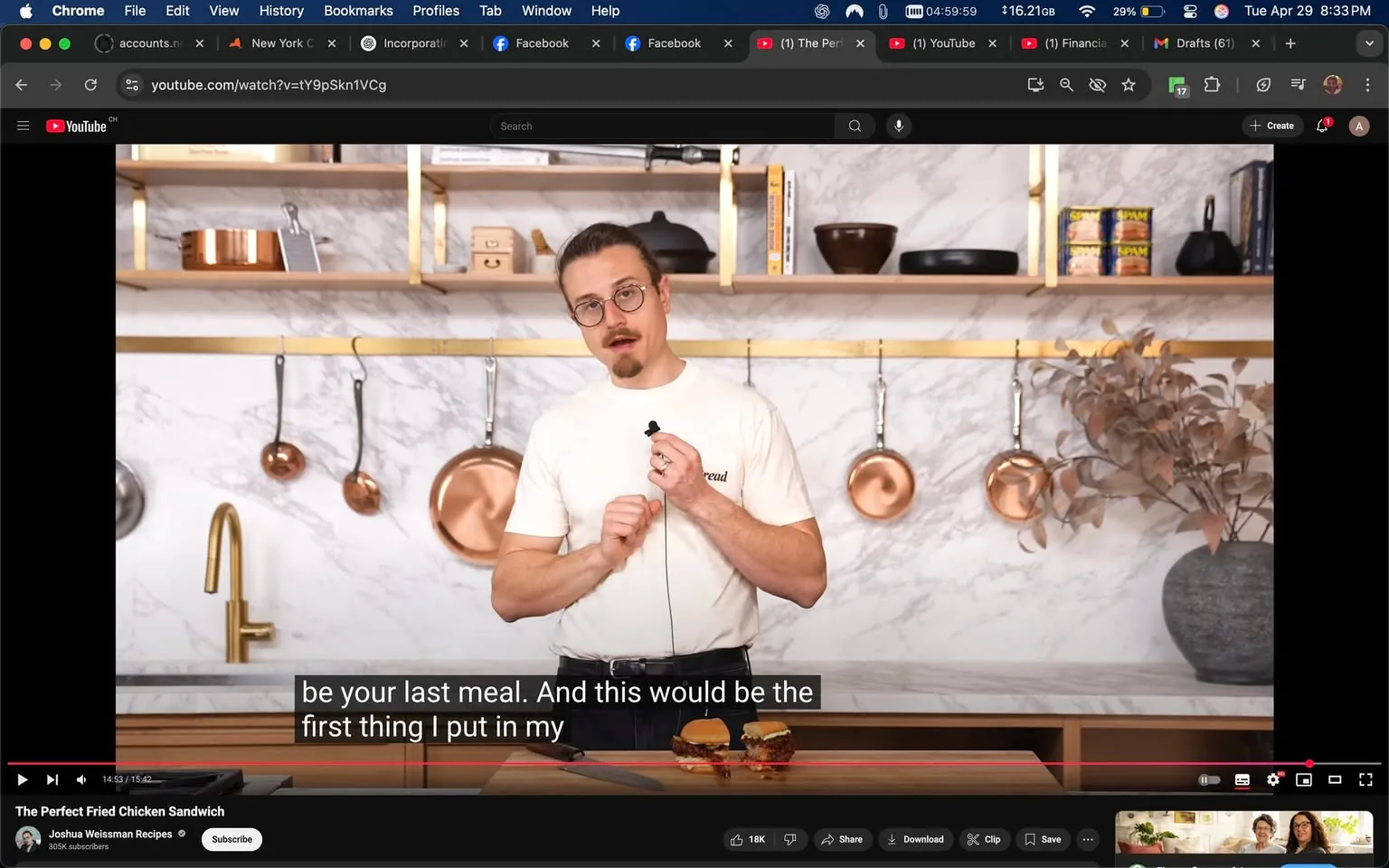 The image features a man standing in a kitchen setting. He has light brown hair tied back in a bun and is wearing round glasses. His shirt is white with the word "bread" printed on it. He appears to be speaking into a microphone, which he holds in his right hand. His left hand is gesturing towards two fried chicken sandwiches placed on a wooden countertop in front of him. 

The sandwiches are stacked one on top of the other, with a golden-brown crust visible on the chicken. The background consists of a marble wall with gold accents, and there are several copper pots and pans hanging on a gold rack above the countertop. A dark ceramic bowl and a can of Spam are also visible on the shelves behind him, along with a small potted plant with dried leaves.

The kitchen has a modern aesthetic, with clean lines and a minimalist design. The lighting appears bright and warm, contributing to a welcoming atmosphere. The man’s expression is focused and engaging, suggesting he is sharing a recipe or cooking tip. 

There are no visible logos or trademarks in the image, and the overall setting is that of a cooking demonstration or tutorial. The video player interface at the bottom indicates that this is a YouTube video, specifically titled "The Perfect Fried Chicken Sandwich," and it shows the channel name "Joshua Weissman Recipes" along with the subscriber count of 305K.