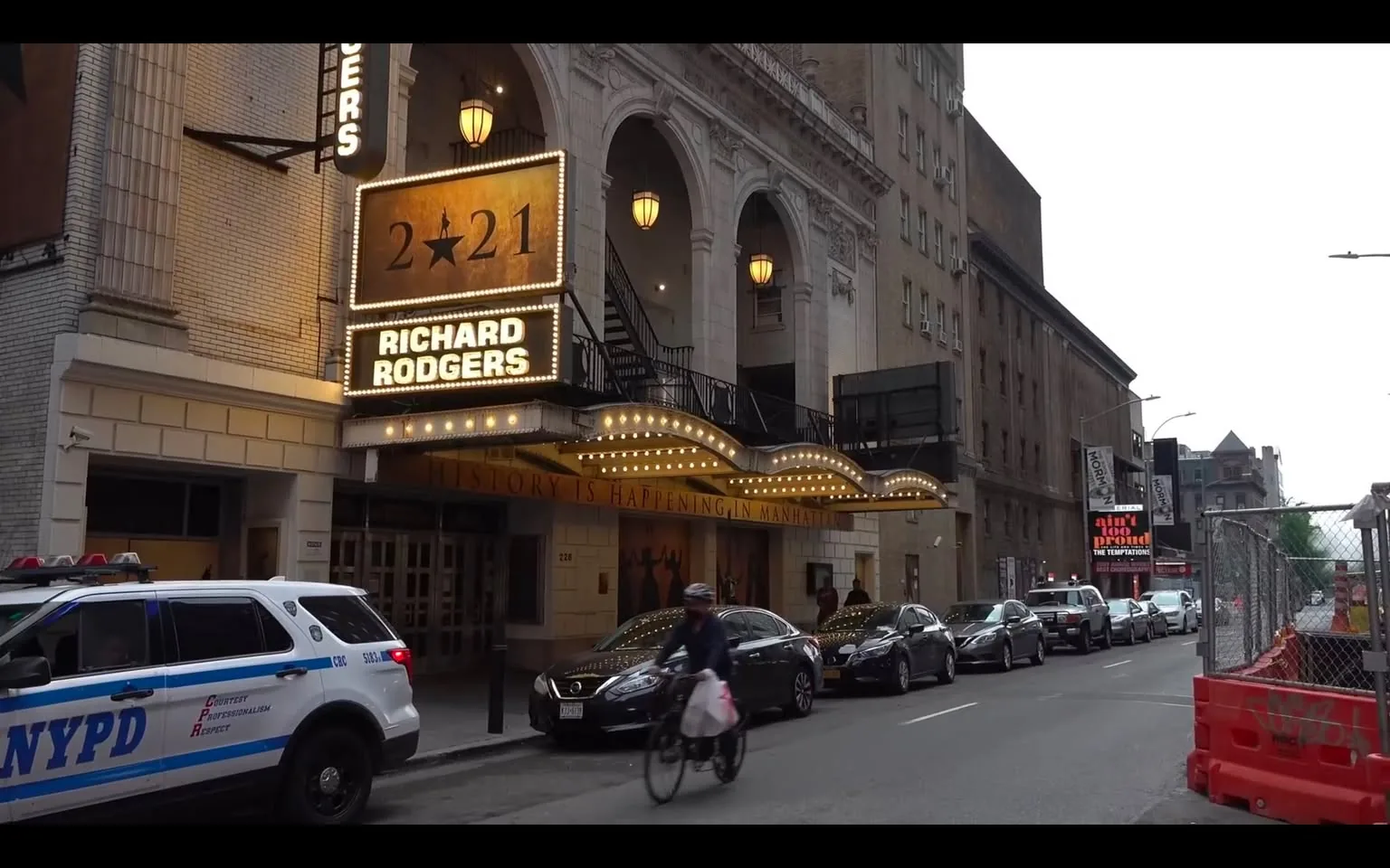 The image depicts the exterior of the Richard Rodgers Theatre, located in a city street setting. The marquee prominently displays the text "2★21" and "RICHARD RODGERS" in bright, illuminated letters. The marquee is adorned with decorative lights, enhancing its visibility. Below the marquee, a sign reads "HISTORY IS HAPPENING IN MANHATTAN," indicating the theater's cultural significance.

The theater's architecture features a grand entrance with arched doorways and decorative elements typical of historic theaters. The facade is primarily white with a textured finish, and there are several hanging light fixtures visible above the entrance.

In the foreground, a police vehicle marked with "NYPD" is parked on the street, indicating a presence of law enforcement in the area. The vehicle has a blue stripe along its side and is equipped with emergency lights. Nearby, a cyclist is seen riding a bicycle, carrying a bag, and wearing a helmet, suggesting an active urban environment.

The street is lined with several parked cars, including a dark sedan and a silver vehicle, indicating a busy area. In the background, there are additional buildings with a mix of architectural styles, some of which appear older and more industrial. A construction site is visible on the right side of the image, marked by orange safety barriers, suggesting ongoing urban development.

The overall atmosphere is that of a bustling city, with a focus on the theater as a cultural landmark. The time of day appears to be early evening, as the lights of the marquee are illuminated against a slightly overcast sky.