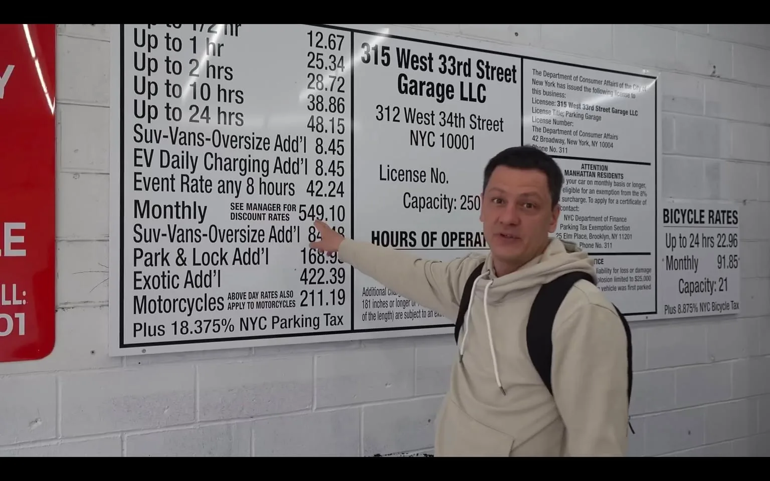 The image depicts a parking garage rate board located at "315 West 33rd Street Garage LLC," with the address "312 West 34th Street, NYC 10001" prominently displayed. The board is white with black text and is divided into sections detailing various parking rates and additional fees. 

At the top, the rates for parking are listed as follows:
- Up to ½ hour: $12.67
- Up to 1 hour: $25.34
- Up to 2 hours: $28.72
- Up to 10 hours: $38.86
- Up to 24 hours: $48.15

Additional fees for larger vehicles are noted:
- SUVs, Vans, Oversized vehicles: $8.45 extra
- EV Daily Charging: $8.45 extra
- Event Rate for any 8 hours: $42.24

The monthly rate is listed as $549.10, with additional charges for oversized vehicles ($8.45), Park & Lock ($168.49), Exotic vehicles ($422.39), and motorcycles ($211.19). A note mentions that these rates are subject to an 18.375% NYC parking tax.

The bottom of the board includes information about the garage's license number and capacity, which is 250 vehicles. There are also hours of operation listed, though they are not fully visible in the image.

To the left of the rate board, there is a red sign that appears to have a phone number, but the text is not fully legible. 

In the foreground, a man is pointing at the rate board. He has short dark hair and is wearing a light-colored hoodie with the hood down. He has a neutral expression and appears to be explaining or discussing the rates. He carries a backpack on one shoulder. 

The background is a concrete wall, typical of a parking garage environment, and the overall setting suggests an indoor parking facility. The lighting is bright, likely fluorescent, illuminating the rate board clearly.