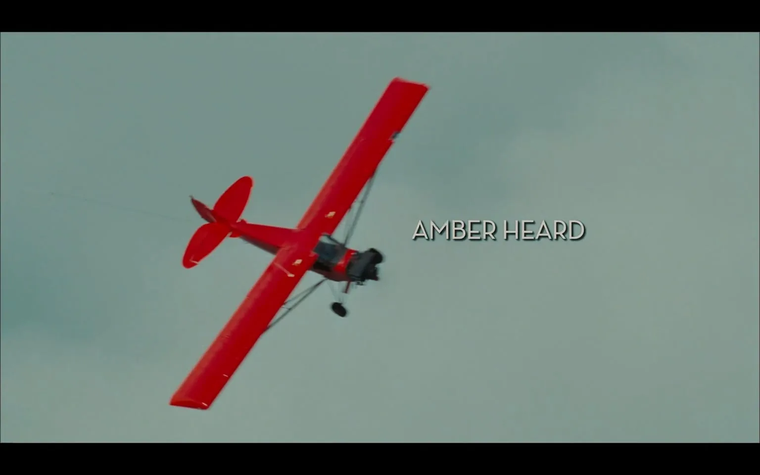 The image features a bright red biplane flying at an angle against a cloudy sky. The aircraft has a classic design, characterized by its open cockpit and large wings. The wings are painted in a vibrant red, and the plane appears to be performing an aerial maneuver, possibly a roll or a loop, as it is tilted to one side. 

In the foreground, the name "AMBER HEARD" is displayed prominently in a bold, white font. The text is positioned centrally on the right side of the image, contrasting against the background and ensuring visibility. The font style is clean and modern, contributing to the cinematic feel of the scene.

The background consists of a pale blue sky with scattered clouds, providing a soft contrast to the vivid red of the plane. The overall atmosphere suggests a sense of adventure and excitement, typical of aerial scenes in films.

No other objects, people, or identifiable landmarks are present in the image. The focus remains solely on the biplane and the text associated with it. The image does not contain any logos, trademarks, or additional text beyond the name displayed.