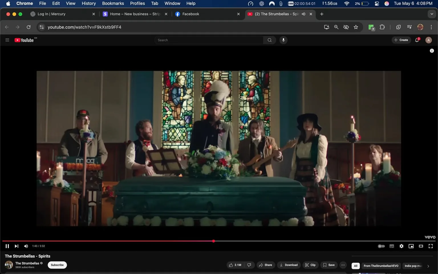 The image depicts a scene from a music video featuring a group of musicians performing in a church-like setting. The focal point is a coffin, which is adorned with a floral arrangement on top, primarily consisting of colorful flowers. The coffin is a teal color and is positioned centrally in the frame.

In the background, there are stained glass windows that depict religious figures, adding to the solemn atmosphere of the setting. The light filtering through the stained glass creates a colorful effect on the walls.

The musicians are dressed in a mix of formal and casual attire, contributing to a whimsical yet respectful vibe. The lead singer, standing at the front, wears a long black coat and a tall hat with a white feather. He appears to be singing or speaking into a microphone. To his left, a man is playing a keyboard, wearing a gray jacket and a cap. Next to him, another musician is playing a guitar, dressed in a brown coat and a bowtie. 

On the right side of the frame, a woman is standing with her hands on her hips, wearing a white blouse and a long skirt, complemented by a black hat. She appears engaged with the performance. Behind her, another musician is playing a guitar, and a woman is seated at a table, possibly observing the performance.

The setting is decorated with candles, some of which are lit, adding to the ambiance. The overall mood of the scene seems to blend elements of a funeral with a musical performance, creating a unique juxtaposition. 

No readable text, logos, or trademarks are visible in the image. The focus remains on the musicians and the coffin, with the stained glass windows providing a colorful backdrop.