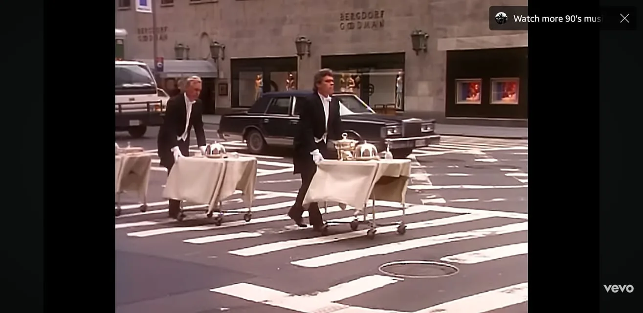 The image depicts two men dressed in formal attire, specifically black tuxedos with white shirts and bow ties, pushing carts across a crosswalk. Each cart is covered with a beige cloth and holds silver serving trays, suggesting they are delivering food or drinks. The man on the left has short, light-colored hair and is wearing white gloves, while the man on the right has darker hair styled in a wavy manner and also wears white gloves. Both men appear focused on their task as they navigate the street.

The background features a city street with a mix of vehicles, including a black sedan and a white truck. The setting appears to be an urban area, likely in a high-end shopping district, as indicated by the presence of a store with the name "Bergdorf Goodman" visible on the building in the background. The architecture is characterized by a stone facade and large windows displaying mannequins and clothing, suggesting a luxury retail environment.

The crosswalk is marked with white stripes, and the scene is bathed in daylight, indicating it is likely daytime. The overall atmosphere seems busy, typical of a metropolitan area, with the men actively engaging in their duties amidst the urban hustle. There are no visible signs, logos, or additional text in the image apart from the store name. The focus remains on the two men and their carts as they traverse the street.