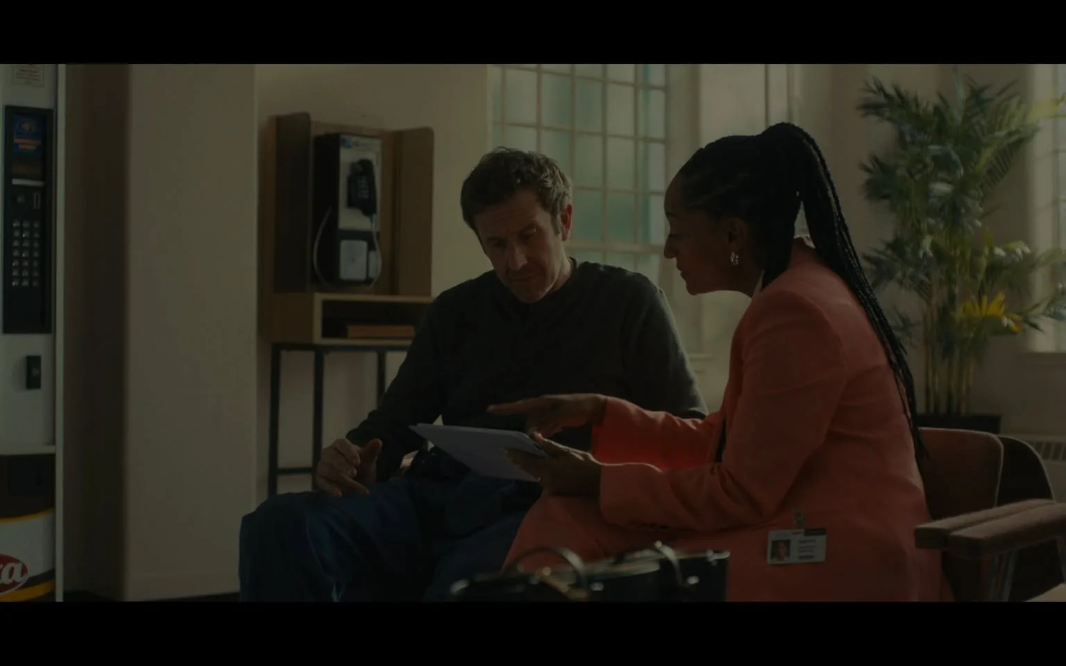 The image depicts two individuals seated in a room that appears to be a waiting area or office space. 

**Setting and Background:**
- The room has a large window with multiple panes, allowing natural light to enter. 
- There is a green plant in the background, adding a touch of greenery to the space.
- To the left, there is a vending machine with a visible logo, likely a beverage brand, and a payphone mounted on the wall behind it.
- The walls are painted a light color, contributing to a bright atmosphere.

**Individuals:**
1. **Person on the Left:**
   - A man with short, slightly wavy dark hair and a medium build.
   - He is wearing a dark gray long-sleeve shirt and blue pants.
   - His expression appears focused as he looks down at a piece of paper held by the woman next to him.
   - He is seated with his hands resting on his knees.

2. **Person on the Right:**
   - A woman with long, braided hair pulled back into a ponytail.
   - She is wearing a light pink blazer over a darker top, giving her a professional appearance.
   - She has a name badge visible on her blazer, which includes a photo and text, though the details are not legible.
   - She is gesturing with her right hand, pointing at the paper in her left hand, which she is showing to the man.

**Actions:**
- The man appears to be engaged in a discussion, possibly reviewing the document the woman is presenting.
- The woman is actively explaining or discussing the content of the paper, indicated by her pointing gesture.

Overall, the scene conveys a professional interaction between the two individuals in a well-lit, informal office environment.