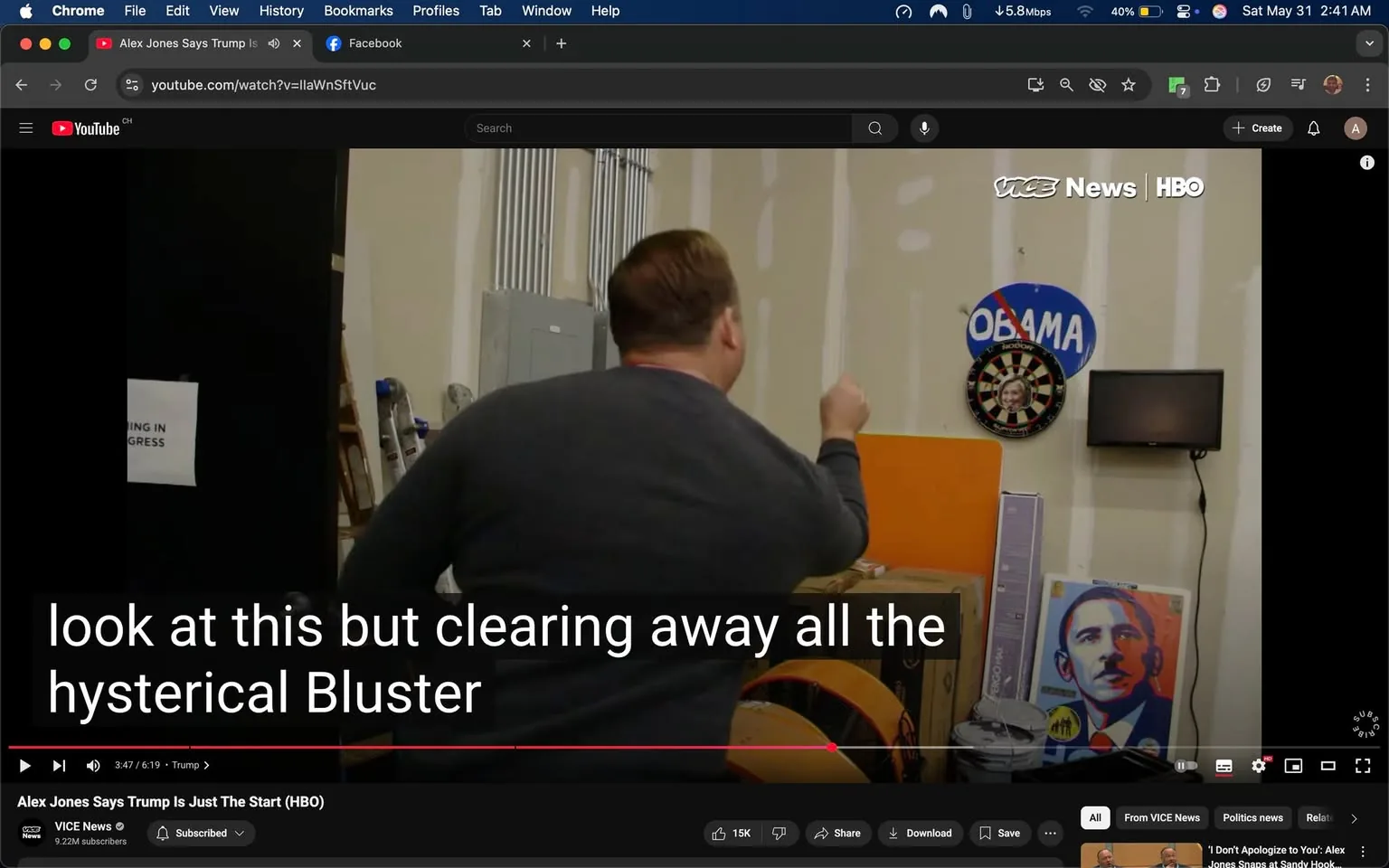 The image depicts a scene from a video featuring a man with short, light brown hair, wearing a dark gray sweater. He is positioned with his back to the camera, gesturing with his right hand as he appears to be speaking or emphasizing a point. The background consists of a partially finished wall, likely in a workspace or studio, with visible drywall and some electrical components. 

On the wall to the right, there is a circular dartboard with a photo of a woman, and above it, a sign that reads "OBAMA" with a red slash through it, indicating disapproval. There is also a flat-screen television mounted on the wall, which is turned off. 

In the foreground, there are several boxes and what appears to be a large, round object, possibly a drum or a similar item, with orange and black colors. A ladder is visible in the background, suggesting ongoing work or renovations in the space. 

The bottom of the image features subtitles that read: "look at this but clearing away all the hysterical Bluster," indicating that the video content may involve commentary or analysis. 

Overall, the setting appears to be informal and somewhat cluttered, typical of a workspace where discussions or presentations are taking place.