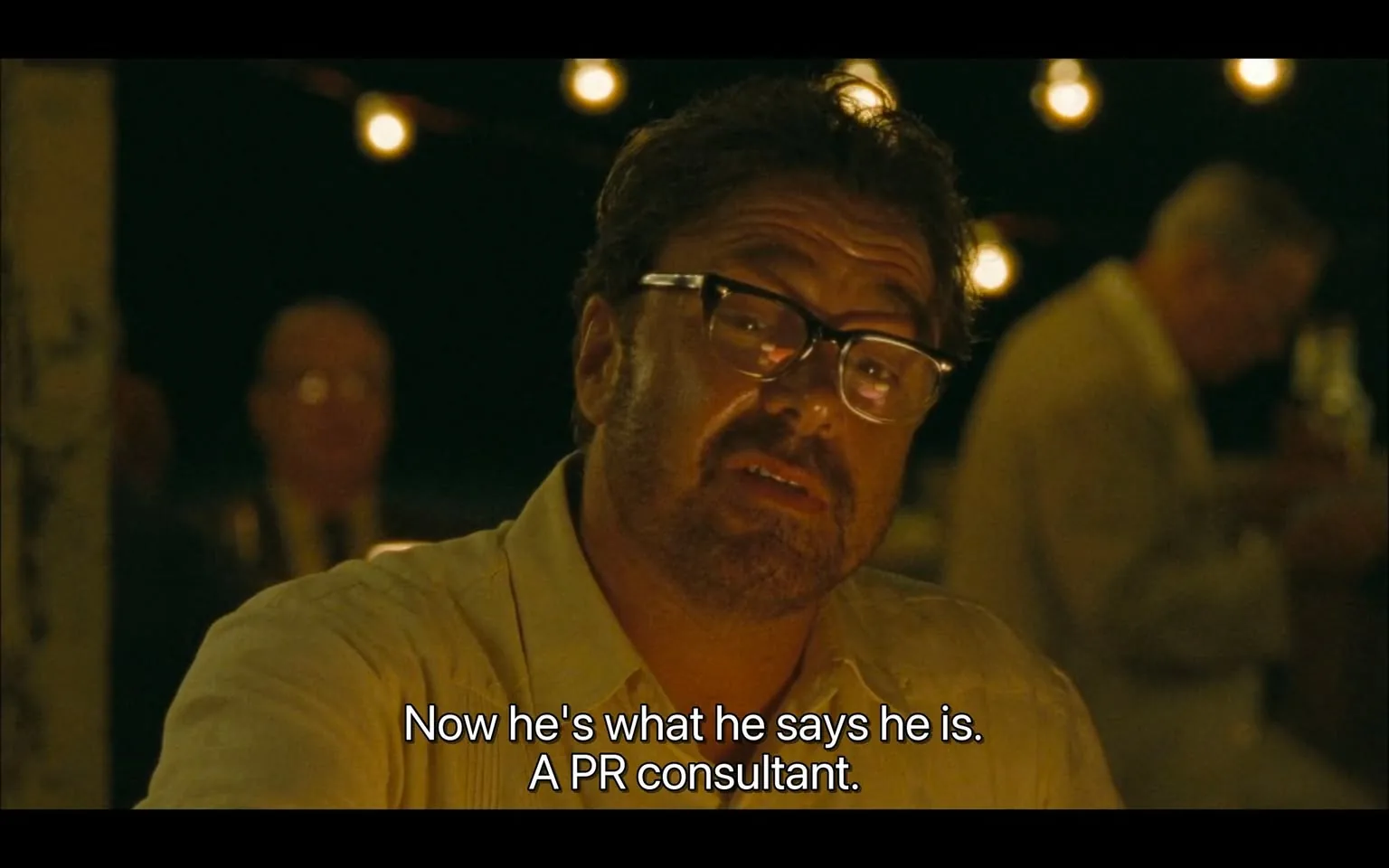 The image depicts a scene from a film, featuring a man speaking directly to the camera. He has a medium build and is wearing a light-colored, short-sleeved shirt. His hair is dark and slightly wavy, and he has facial hair, specifically a beard and mustache. He is also wearing rectangular glasses with a thick frame. The man appears to be in a conversational or storytelling mode, with a serious expression on his face.

In the background, there are several indistinct figures, suggesting that the setting is a gathering or dinner party. The lighting is warm and ambient, with soft, glowing string lights visible, creating a cozy atmosphere. The background is somewhat blurred, but it appears to be outdoors, possibly in a garden or patio area, given the presence of greenery and the informal setting.

The text displayed at the bottom of the image reads: "Now he's what he says he is. A PR consultant." This suggests that the dialogue is part of a narrative discussing someone's profession or identity.

Overall, the scene conveys a sense of intimacy and engagement, with the man at the forefront clearly the focus of the moment. The combination of his attire, the lighting, and the background elements contribute to a relaxed yet serious ambiance typical of a personal conversation.