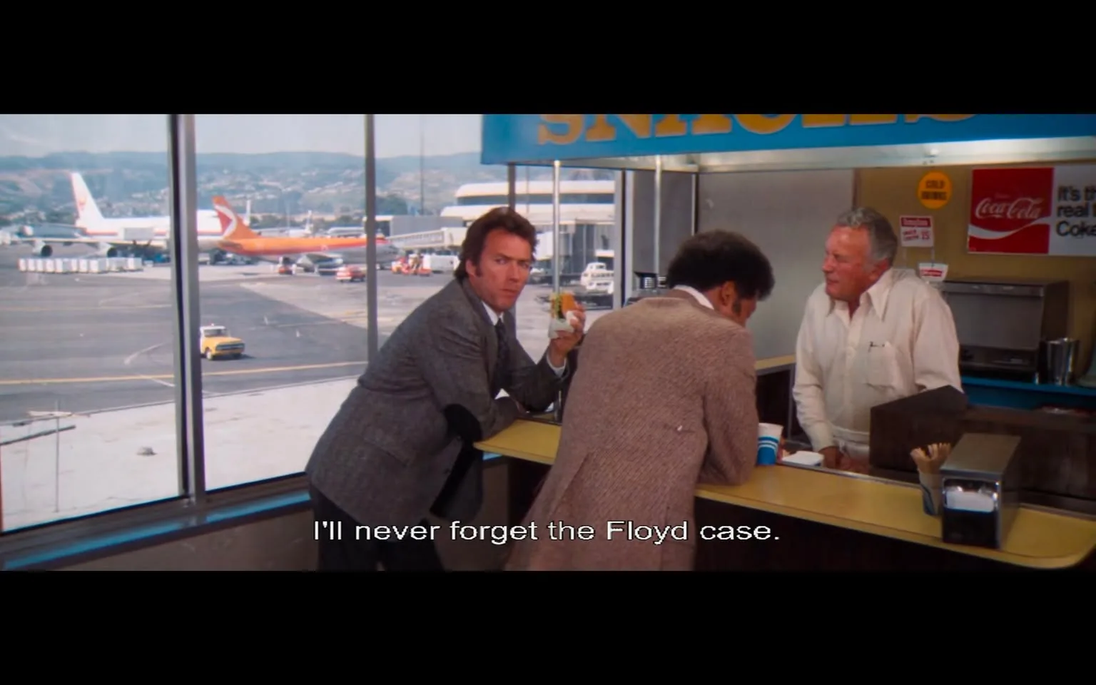 The image depicts a scene set in an airport terminal, specifically at a food counter. The counter has a sign that reads "SNACKS" in large, bold letters, with a bright blue background. Behind the counter, there is a Coca-Cola advertisement that states, "It's the real thing," along with a logo. 

In the foreground, three men are engaged in conversation. The man on the left is wearing a light gray suit with a white shirt and a tie. He has short, slightly tousled hair and is holding a drink in one hand while looking back at the camera with a serious expression. He appears to be in mid-conversation, possibly sharing a story or making a point.

The man in the middle has curly black hair and is wearing a brown, textured jacket. He is facing the counter, leaning slightly forward, and appears to be listening intently to the man on the right. The man on the right is older, with gray hair and a light-colored shirt. He is standing behind the counter, looking at the middle man with a friendly demeanor, possibly engaging in a light-hearted discussion.

In the background, large windows reveal a view of the airport tarmac, where several airplanes are parked. Notably, there is an orange and white aircraft visible, along with other planes and ground vehicles. The landscape beyond the airport shows rolling hills under a clear sky.

Overall, the scene captures a casual yet engaging moment among the three men, set against the bustling backdrop of an airport environment.