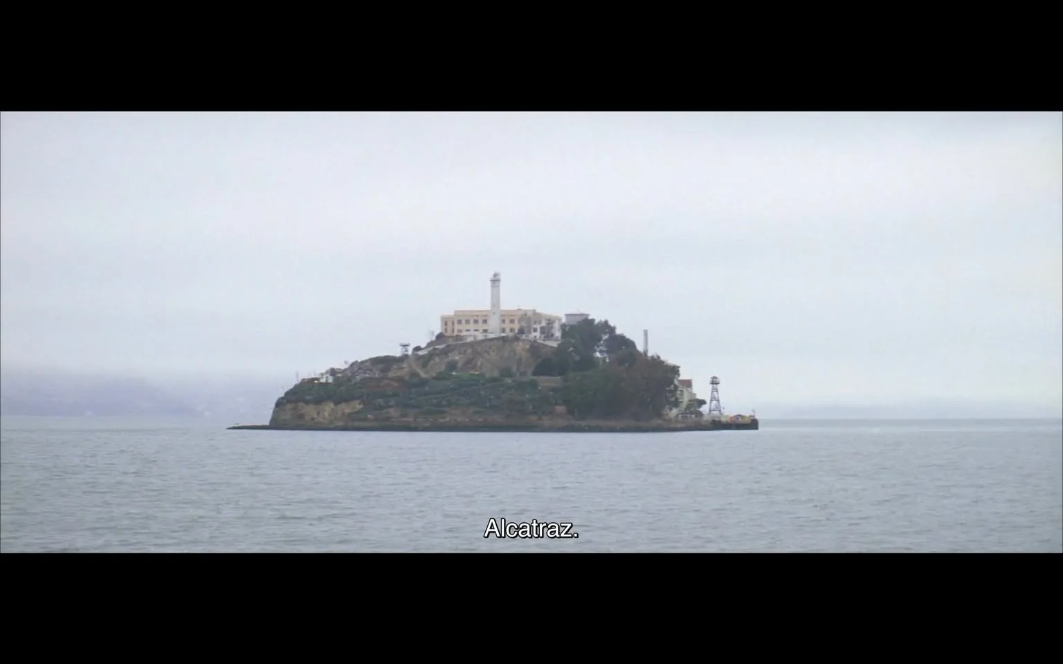 The image depicts a distant view of Alcatraz Island, situated in the water. The island features a prominent building at the top, which appears to be a historical structure, likely the former prison, characterized by its rectangular shape and multiple windows. A tall lighthouse is visible, standing to the left of the main building, with a white cylindrical design that extends upward. 

Surrounding the main structure are patches of greenery, indicating some vegetation on the island. The shoreline of the island is rocky and uneven, with a slight incline leading up to the buildings. To the right, there is a small structure resembling a lighthouse or lookout tower, painted in white and possibly used for navigation or observation.

The water surrounding the island is calm, reflecting the overcast sky, which is filled with gray clouds, suggesting a cloudy day. The overall atmosphere appears serene, with no visible human activity on the island. 

At the bottom of the image, there is a subtitle that reads "Alcatraz," indicating the name of the island. The text is centered and in a simple, sans-serif font, contrasting with the background. The image captures a historical and iconic location, known for its past as a federal prison.