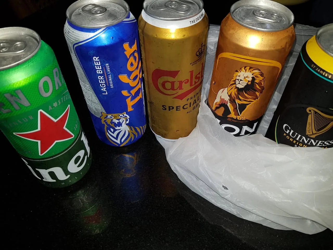 The image displays five cans of beer arranged closely together on a dark surface, likely a table. The cans are positioned in a way that allows for clear visibility of their labels and designs.

1. **Heineken**: The first can on the left is green with a prominent red star logo. The text on the can reads "Heineken" and "Malt Lager" in white lettering. The can has a textured surface, typical of Heineken's branding.

2. **Tiger Beer**: Next to Heineken is a blue can labeled "Tiger" with the words "Lager Beer" and "Original Lager" prominently displayed. The can features a graphic of a tiger, which is part of its branding.

3. **Carlsberg**: The third can is gold and white, labeled "Carlsberg" with "Premium Lager" and "Special Brew" in red text. The can has a crown logo above the brand name, indicating its premium status.

4. **Lion Stout**: The fourth can is a darker gold with a graphic of a lion on it. The label reads "Lion" and "Stout" with "Wood" below the lion image. The design suggests a strong, bold beer.

5. **Guinness**: The last can on the right is black with a gold logo. It reads "Guinness" and "Foreign Extra Stout" in white text. The can features the iconic harp logo associated with the brand.

The cans are partially obscured by a white plastic bag, which is crumpled and appears to be resting on the surface. The background is dark, enhancing the visibility of the colorful cans. There are no people or additional objects in the image, focusing solely on the beer cans. The overall setting appears casual, possibly indicating a gathering or personal selection of beverages.