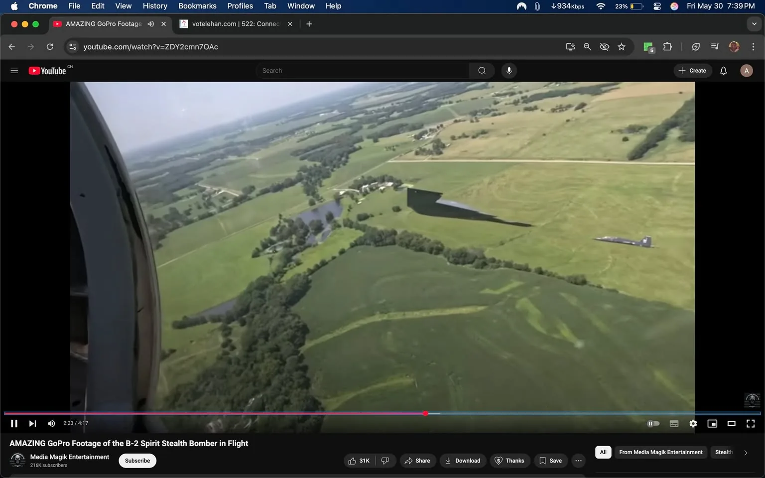 The image shows a view from an aircraft, likely a military plane, as it flies over a rural landscape. The perspective is from the side of the aircraft, capturing the wing and part of the fuselage. The wing appears to be large and has a sleek, aerodynamic design, typical of stealth bombers.

Below, the landscape consists of expansive green fields, interspersed with patches of trees and a winding river. The fields are divided into various shades of green, indicating different types of vegetation or farming practices. There are also some darker areas, possibly indicating water bodies or shaded regions from trees.

In the distance, there are more fields and a few structures, likely farmhouses or barns, visible among the greenery. The sky is mostly clear with a few clouds, suggesting a bright day. The overall setting appears to be rural, with a mix of agricultural land and natural features.

No readable text, logos, or identifiable brands are present in the image. The focus is primarily on the aerial view of the landscape and the aircraft's wing. The image captures a moment in flight, showcasing the vastness of the countryside below.