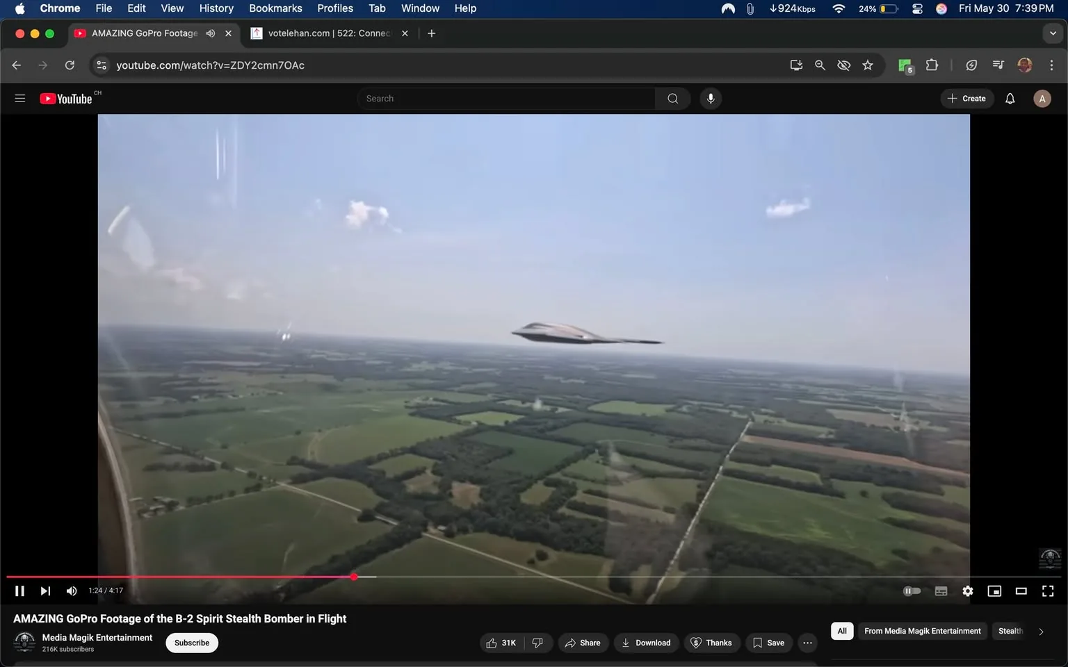 The image shows a video still from a YouTube clip titled "AMAZING GoPro Footage of the B-2 Spirit Stealth Bomber in Flight." The video features a view from an aircraft, capturing the B-2 Spirit stealth bomber flying in the sky. 

In the foreground, the B-2 bomber is prominently displayed, characterized by its distinctive flying wing design. The aircraft has a sleek, dark exterior with a smooth surface, and it appears to be flying at a low altitude over a vast landscape. The bomber is positioned slightly to the right of the frame, angled as if it is in motion.

The background consists of a wide expanse of green fields and patches of trees, indicating a rural or agricultural area. The terrain is divided into various sections, possibly indicating different types of crops or land use. There are some clouds in the sky, which is mostly clear, suggesting a bright day.

In the lower part of the image, there is a YouTube video player interface visible, including a red progress bar indicating the video is at 1 minute and 24 seconds into a total length of 4 minutes and 17 seconds. Below the video, there are various buttons such as "Like," "Share," "Download," and "Thanks," along with a subscriber count of 216K for the channel "Media Magik Entertainment."

No people are visible in the image, and there are no readable texts or logos apart from the YouTube interface. The overall setting suggests an aviation or military context, focusing on the capabilities of the B-2 bomber.