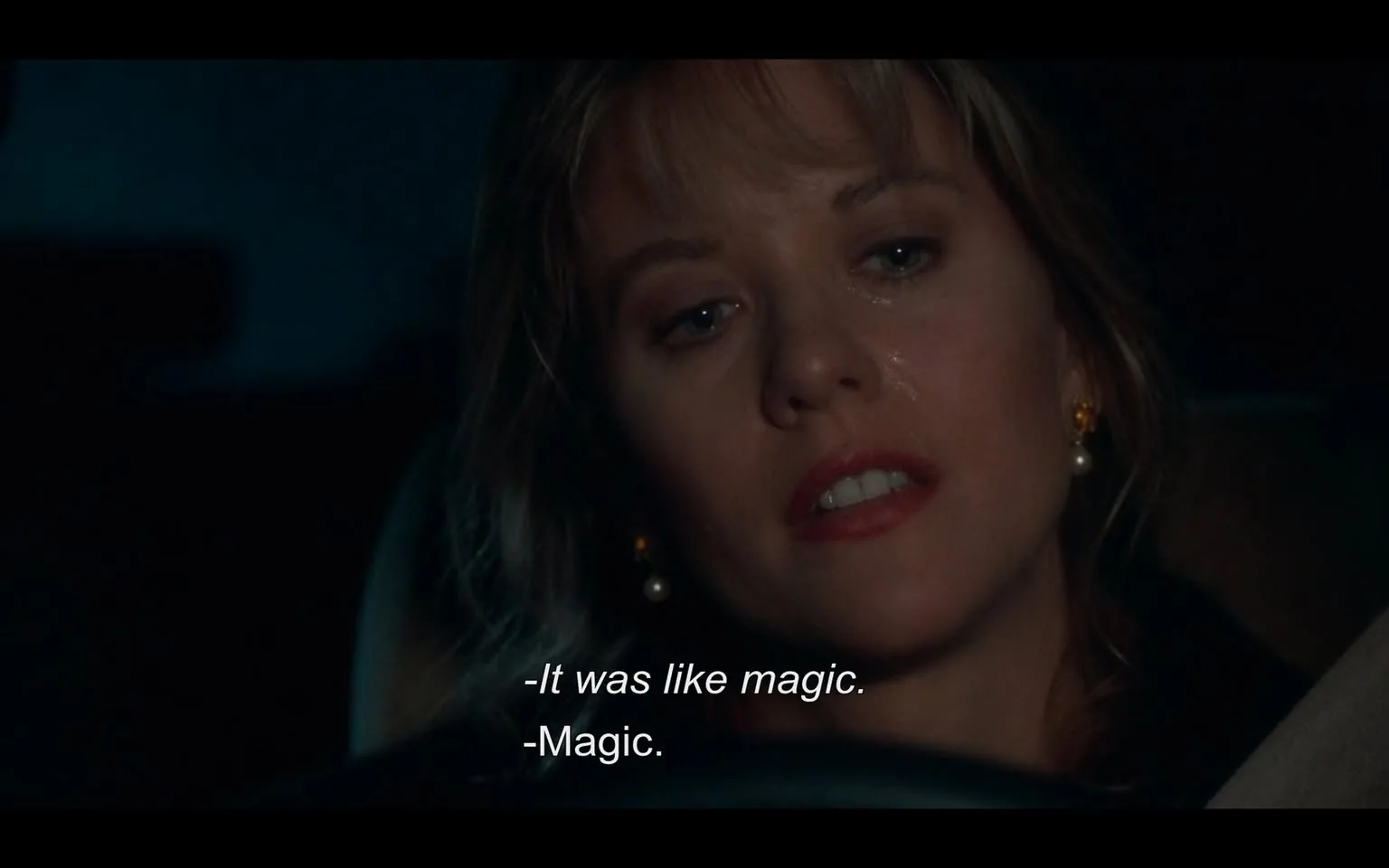 The image depicts a close-up of a woman sitting in a car, with a dark background suggesting nighttime. She has light brown hair that appears slightly wavy and is styled with some bangs. Her facial expression shows a mix of contemplation and emotion, with her eyes slightly glistening, possibly indicating tears. She has a subtle sheen on her cheeks, which may suggest she has been crying or is emotional. 

The woman is wearing small, round pearl earrings that stand out against her skin. Her lips are painted a soft shade of red, and her makeup is understated, enhancing her natural features. She is dressed in a dark-colored top, which is not fully visible due to the angle of the shot.

In the foreground, part of the car's interior is visible, including a section of the steering wheel and the dashboard, which is mostly out of focus. The lighting in the car is dim, with a blueish tint that adds to the nighttime atmosphere. 

Text is visible at the bottom of the image, indicating dialogue: "-It was like magic." and "-Magic." The text is white and clearly legible against the darker background. 

The overall setting suggests a private moment, likely a conversation taking place in the car, but no specific location or external environment is visible beyond the interior of the vehicle. There are no identifiable brands, logos, or trademarks present in the image.