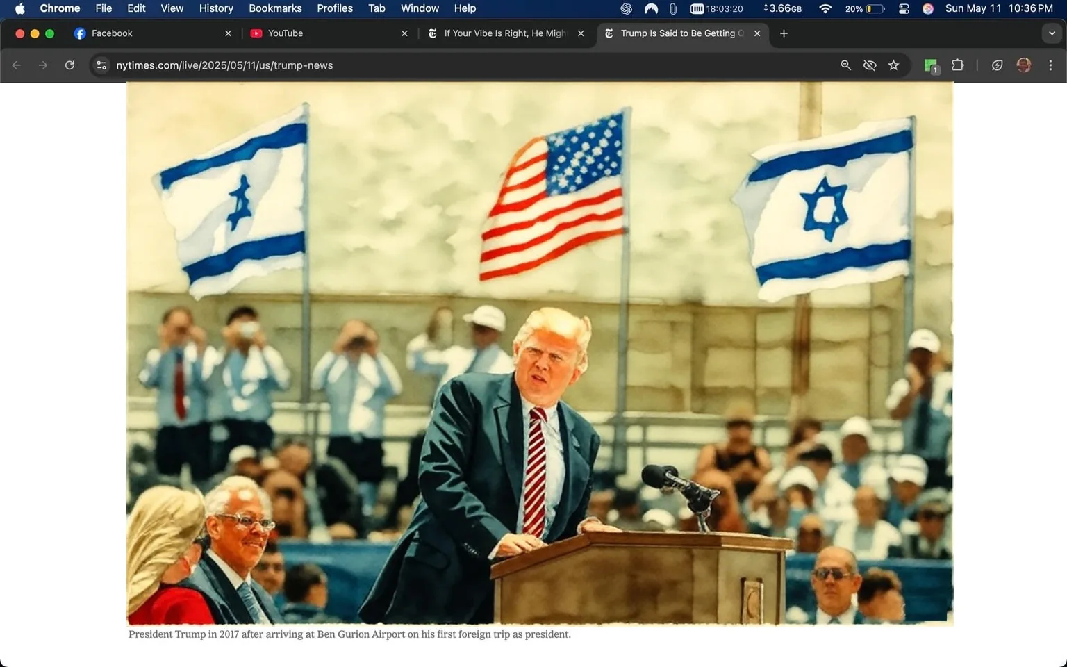 The image depicts a scene from a public event featuring a man at a podium, who appears to be delivering a speech. He has light-colored hair and is wearing a dark suit with a white shirt and a red and blue striped tie. The podium is wooden and has a microphone on it. In the background, there are several flags visible, including the American flag and the Israeli flag, which indicates that the event may be taking place in Israel, specifically at Ben Gurion Airport.

To the left of the podium, there is a woman with long, wavy blonde hair, wearing sunglasses and a red dress. She appears to be looking at the speaker. To the right, there is a man with gray hair, dressed in a suit and tie, who is smiling and looking at the speaker as well. Behind them, a crowd of people is visible, many of whom are wearing white shirts and are holding cameras or smartphones, seemingly capturing the moment.

The setting appears to be outdoors, with a bright, sunny atmosphere. The background features a blurred view of a building, suggesting an airport environment. The flags are waving gently in the breeze, adding to the lively ambiance of the event. The overall scene conveys a sense of formality and significance, likely related to a political or diplomatic occasion.