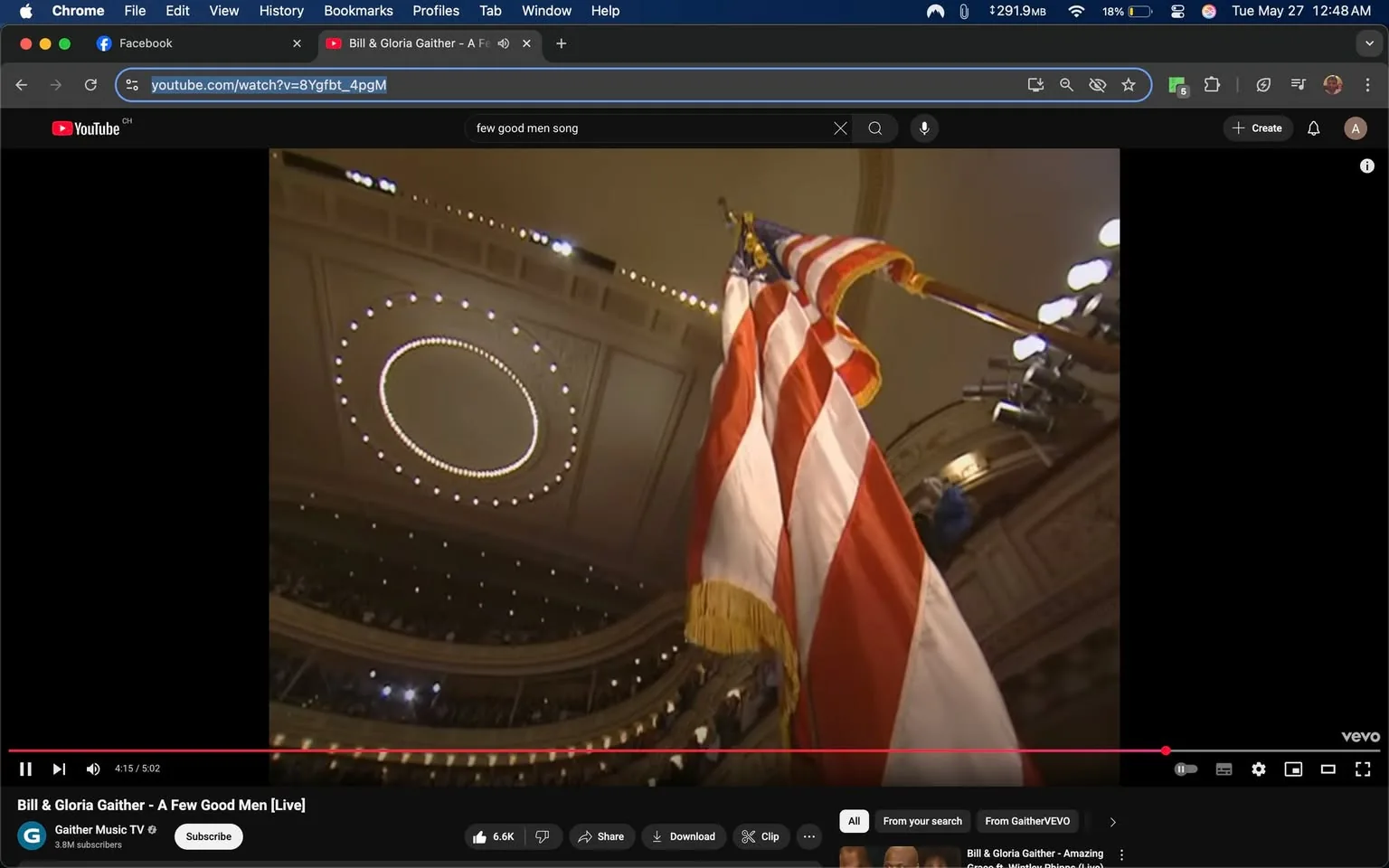 The image shows a low-angle view of an American flag prominently displayed in a large indoor venue. The flag features red and white stripes with a blue field containing white stars, and it is mounted on a pole with a gold fringe at the bottom. The setting appears to be a grand hall or auditorium, characterized by ornate architectural details. 

In the background, the ceiling is visible, featuring a circular light fixture composed of small lights arranged in a circular pattern. The ceiling has intricate designs, suggesting a historic or formal venue. The walls of the auditorium are lined with balconies, indicating that it is designed to accommodate a large audience. 

The lighting in the venue is warm, with several overhead lights illuminating the space. There are also visible audience members in the background, though they are not clearly defined due to the angle and focus on the flag. 

The video player interface at the bottom of the image shows that the video is titled "Bill & Gloria Gaither - A Few Good Men [Live]," indicating that this is a performance or musical event. The video is currently at 4 minutes and 15 seconds into a total duration of 5 minutes and 2 seconds. The YouTube logo is visible in the top left corner, along with the channel name "Gaither Music TV," which has 3.8 million subscribers. 

Overall, the image captures a moment during a live performance in a formal setting, highlighting the American flag as a central visual element.