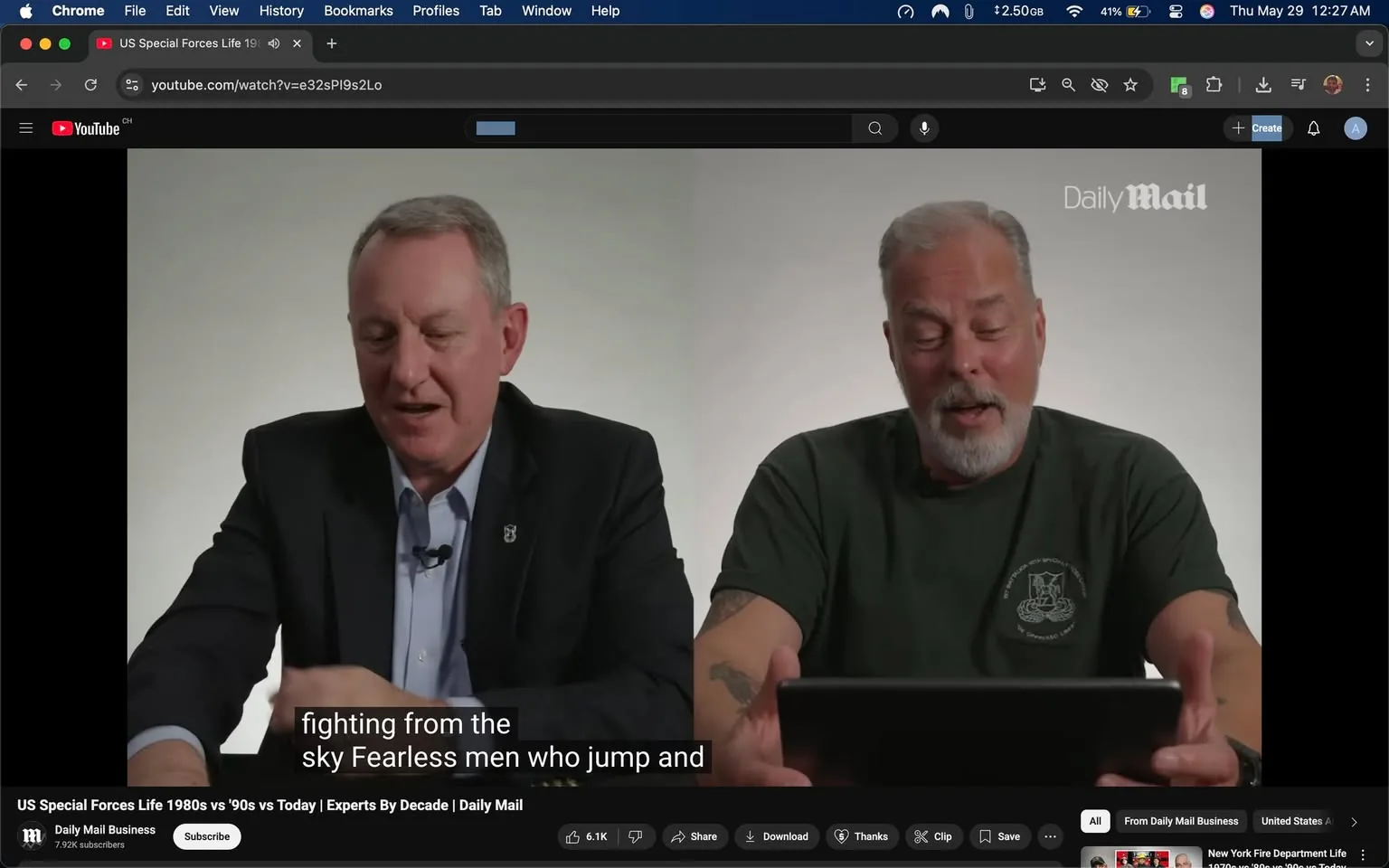The image features two men seated side by side, engaged in a discussion. The background is a plain, light-colored wall, creating a neutral setting that focuses attention on the speakers.

On the left, the first man is wearing a black suit jacket over a light blue dress shirt. He has short, neatly combed hair and appears to be in his mid-50s. His expression is friendly and engaged as he speaks. He has a microphone clipped to his shirt and is gesturing with his right hand while holding a tablet in his left hand.

The second man, on the right, is dressed in a dark green t-shirt with a logo or emblem on the left side, which is not clearly visible. He has a beard that is mostly gray, and his hair is also gray and short. He appears to be slightly older than the first man, possibly in his late 50s or early 60s. His expression is animated, and he is looking at the tablet in front of him, possibly reading or referencing it while speaking.

Above the two men, there is a logo that reads "Daily Mail," indicating the source of the video. The bottom of the image includes a video player interface with various controls, including options to share, download, and save the video. There are also visible viewer statistics, such as "6.1K" and "7.92K subscribers," suggesting the video has garnered a significant amount of attention.

The text visible on the screen reads, "fighting from the sky Fearless men who jump and," indicating the topic of their discussion, which seems to relate to military or special forces operations. The overall atmosphere of the image is professional, likely intended for an informative or educational video.