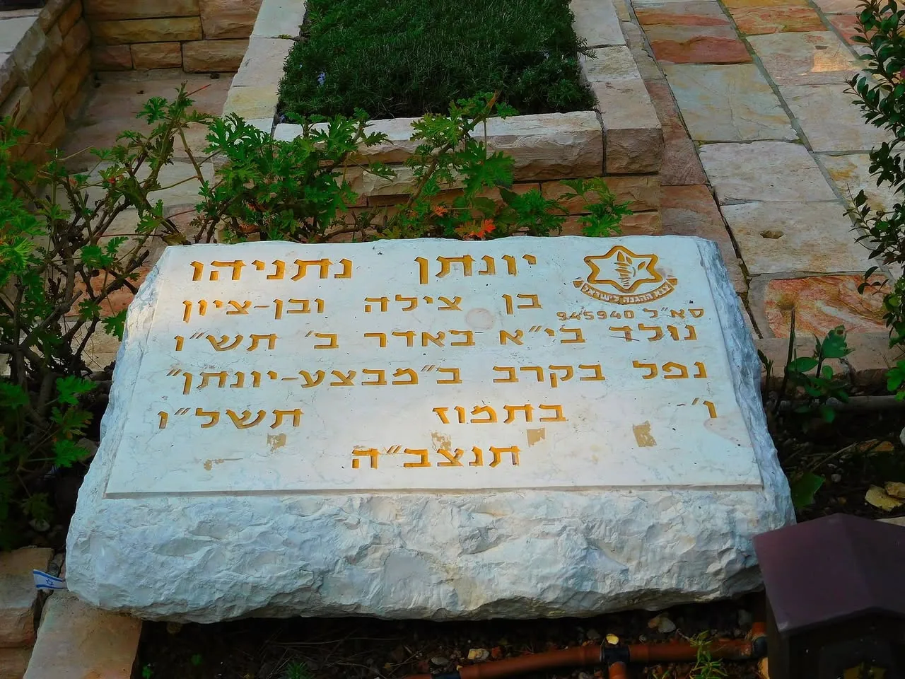 The image features a large, rectangular stone monument set in a landscaped area. The stone is predominantly white with a rough texture and appears to be made of natural rock. It is positioned on a pathway made of stone tiles, which are arranged in a pattern and have a warm, earthy color palette, including shades of beige and light brown.

On the stone, there is engraved text in Hebrew, which is prominently displayed in a gold color. The text is arranged in multiple lines and includes the name "יונתן נתניהו" (Yonatan Netanyahu) at the top, followed by additional inscriptions that commemorate his life and service. The engraving is clear, with the letters being well-defined against the stone's surface.

To the left of the stone, there are green plants and shrubs, adding to the natural setting. The background features more stonework, possibly part of a larger garden or memorial area. The overall environment appears serene and well-maintained, suggesting it is a place of remembrance or honor.

There are no people visible in the image, and the focus remains solely on the stone monument and its surroundings. The lighting appears to be natural, indicating that the photo may have been taken during the day. The scene conveys a sense of respect and tranquility, typical of memorial sites.