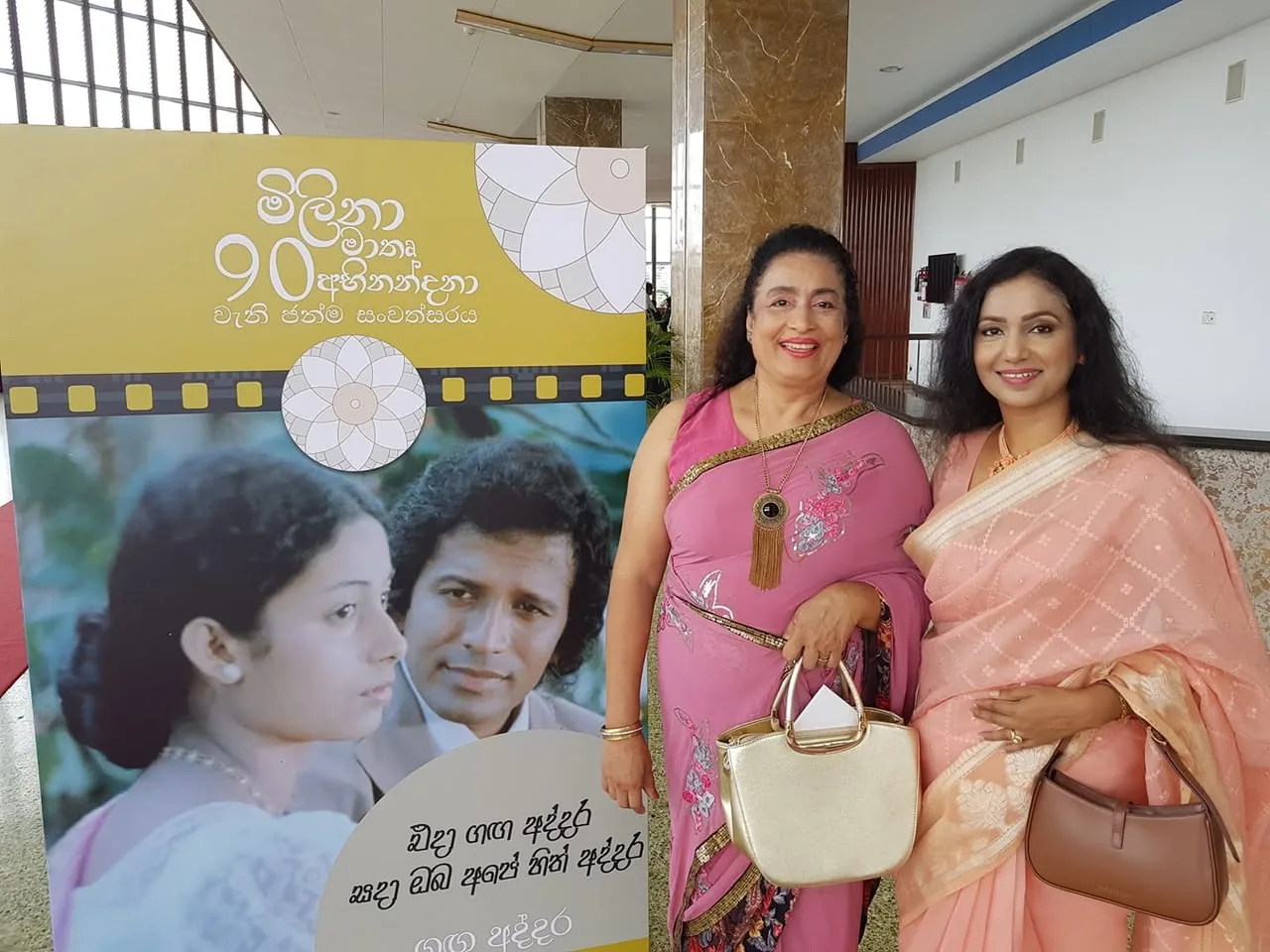 The image features two women standing in front of a large promotional display. The display has a yellow background with text in Sinhala, which translates to "Milana 90th Anniversary." Below the title, there are images of two individuals, a man and a woman, likely from a film or television show, with the man wearing a suit and the woman in a traditional outfit.

The woman on the left is wearing a vibrant pink saree adorned with intricate silver embroidery. She has long, dark hair styled in loose waves and is smiling broadly, displaying a cheerful expression. Around her neck, she wears a long necklace with a large pendant. In her right hand, she holds a gold handbag.

The woman on the right is dressed in a soft pink saree with a subtle pattern, complemented by a light-colored blouse. Her hair is also long and wavy, and she has a gentle smile on her face. She is holding a brown handbag in her left hand, which has a structured design.

The setting appears to be an indoor venue, possibly for an event or celebration, with large windows in the background allowing natural light to filter in. The floor is tiled, and there are decorative elements in the background, including a marble wall. The overall atmosphere suggests a formal or celebratory occasion, likely related to the film industry, given the context of the display.