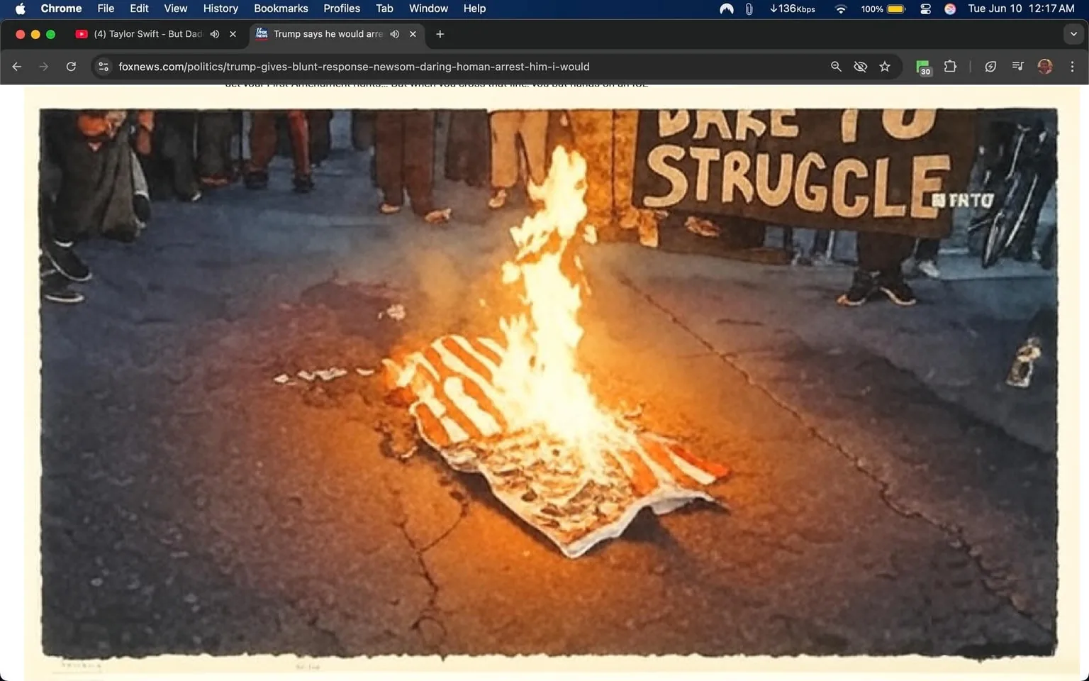The image depicts a scene of protest in Los Angeles, where a group of individuals is gathered around a burning American flag. The flag is laid flat on the ground and is engulfed in flames, with bright orange and yellow fire contrasting against the darker asphalt surface. The flag appears to be partially charred, with some sections still intact while others are consumed by the fire.

In the background, there are several individuals standing and observing the burning flag. They are dressed in casual clothing, including t-shirts and jeans, but specific details about their clothing or features are not discernible due to the focus on the flag and fire. Some individuals are holding signs, one of which is partially visible and reads "DARE TO STRUGGLE." The sign is black with white text, and it appears to be made of cardboard or a similar material.

The setting is an outdoor urban environment, likely a street or public area, as indicated by the asphalt surface and the presence of a crowd. The lighting suggests it may be evening or nighttime, as the flames provide the primary source of illumination in the image.

No logos, trademarks, or identifiable brands are visible in the image aside from the text on the sign. The overall atmosphere appears to be one of protest, with the act of burning the flag symbolizing a strong political statement. The image captures a moment of tension and activism, reflecting the ongoing discourse surrounding immigration and law enforcement in the context of the protests mentioned in the accompanying text.