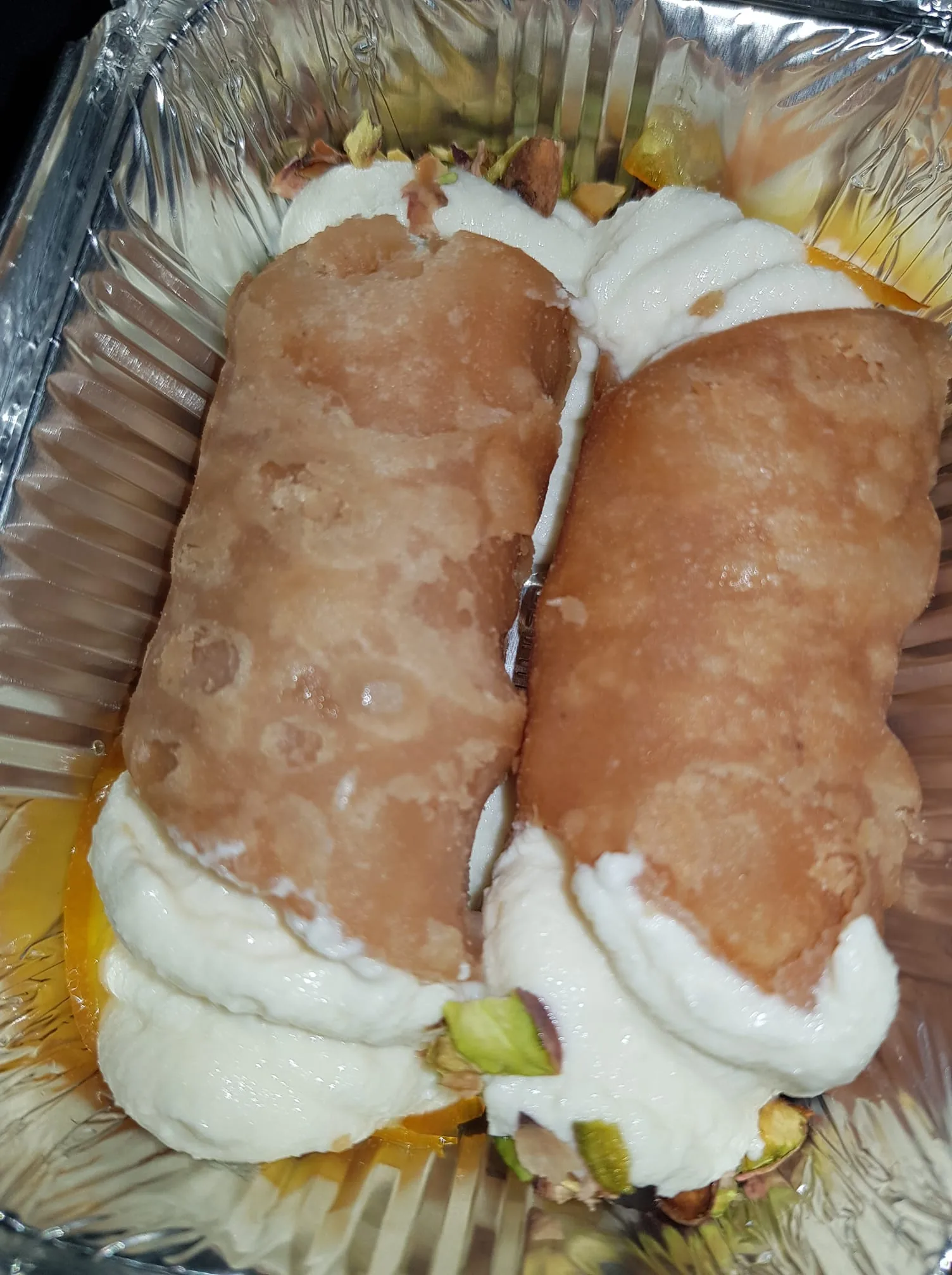 The image shows two cylindrical desserts placed in a metallic foil container. Each dessert appears to be a type of pastry, possibly resembling cannoli or similar items. The outer layer of the pastries is a light brown color with a slightly crispy texture, featuring a smooth surface with some irregularities and small bubbles, indicating a fried or baked preparation.

Inside the pastries, there is a creamy filling that is predominantly white, suggesting a rich, sweet cream or ricotta-like substance. The filling is visible at both ends of the pastries, spilling slightly over the edges. Scattered among the filling are small pieces of chopped pistachios, adding a pop of green color and texture.

Beneath the pastries, there is a layer of what appears to be a syrup or sauce, possibly orange in color, which may be a fruit-based syrup or a sweet glaze. The container itself is rectangular, with a shiny metallic interior and ridged sides, typical of takeout or dessert packaging.

Overall, the image focuses solely on the desserts without any visible text, branding, or additional context that would indicate a specific location or event. There are no people or other objects present in the frame, emphasizing the food items as the main subject.