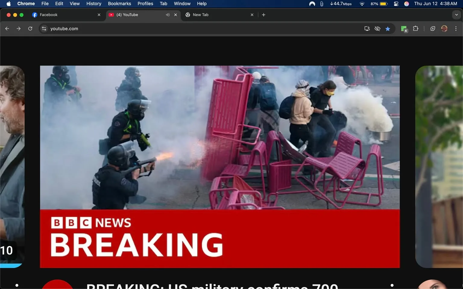 The image depicts a chaotic scene, likely from a protest or riot. In the foreground, several individuals are engaged in a tense situation, with smoke filling the air. 

1. **Text and Logos**: At the bottom of the image, there is a prominent red banner with white text that reads "BBC NEWS BREAKING." Below this, there is additional text stating, "BREAKING: US military confirms 700." 

2. **People and Clothing**: 
   - In the center-left, a person in a black uniform, possibly a police officer, is aiming a firearm that appears to be discharging. They are wearing a helmet and a face shield, with a green harness visible on their uniform.
   - To the right of this individual, another person is seen wearing a beige hoodie and a backpack, appearing to be in motion. They have their head turned, possibly looking back at the unfolding situation.
   - A third individual, dressed in a black jacket and wearing a beanie, is also moving away from the scene, suggesting a sense of urgency.
   - The clothing of the individuals varies, with some wearing casual attire like hoodies and backpacks, while others are in tactical gear.

3. **Setting and Background**: The background is filled with smoke, which may indicate the use of tear gas or other crowd control measures. Scattered around the scene are several pink plastic chairs, some overturned, suggesting a struggle or a hasty retreat. The ground appears to be asphalt, typical of an urban environment.

4. **Environmental Context**: The overall atmosphere is tense and chaotic, indicative of a confrontation between law enforcement and protesters. The presence of smoke and the actions of the individuals suggest a high-stress situation, likely related to civil unrest.

5. **Additional Elements**: The image captures a moment of action, with movement and urgency evident among the individuals present. The smoke adds a layer of obscurity, further emphasizing the chaotic nature of the scene.

Overall, the image conveys a significant moment of conflict, with clear indicators of a protest or riot situation, marked by the presence of law enforcement and civilians in a state of distress.
