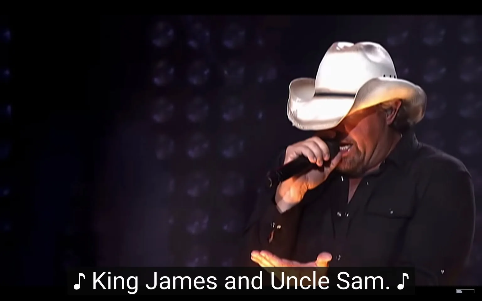 The image depicts a male performer singing on stage. He is wearing a white cowboy hat that has a wide brim and a slightly curved top. His hair is light-colored and appears to be short. The performer is dressed in a black button-up shirt, which is unbuttoned at the collar, revealing a hint of his neck. He is holding a microphone in his right hand, positioned close to his mouth, while his left hand is gesturing expressively, with fingers slightly curled.

The background features a dark setting with a gradient of colors, primarily black, and hints of purple or blue lights that seem to be emanating from the stage. There are circular lights or reflections visible in the background, creating a dynamic atmosphere typical of a live performance. 

At the bottom of the image, there is readable text that states, "♪ King James and Uncle Sam. ♪" This text appears to be part of the lyrics or a caption related to the performance. 

The overall setting suggests a concert or live music event, with the focus on the performer as he engages with the audience. The lighting is dramatic, enhancing the visual impact of the scene. The performer’s expression is not fully visible due to the angle of the hat, but he appears to be focused on his performance. 

No identifiable logos, brands, or trademarks are present in the image. The context is clearly that of a musical performance, but no specific details about the event or location are provided.