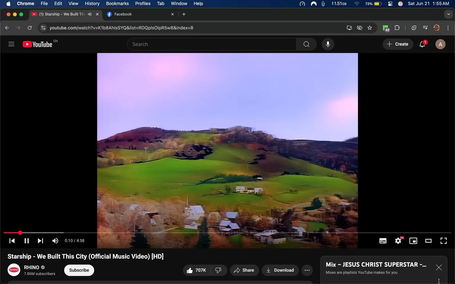 The image displays a still from the official music video for "We Built This City" by Starship. The video is playing on a YouTube interface, which is visible on the screen. 

In the foreground, the video features a landscape scene characterized by rolling green hills. The hills are interspersed with patches of brown and orange foliage, suggesting a transition between seasons, possibly autumn. There are several white and gray buildings visible at the base of the hills, likely homes or small structures, with some trees scattered throughout the area. The sky above is a gradient of soft colors, transitioning from pale blue to light pink, indicating either dawn or dusk.

The YouTube interface includes a black sidebar on the left, which is mostly empty, and a red progress bar at the bottom indicating the video is currently at 10 seconds out of a total length of 4 minutes and 58 seconds. Below the video, there is a channel name "RHINO" with a subscriber count of 7.86 million. The options to "Subscribe," "Share," and "Download" are also visible.

No people are present in the image, and there are no discernible facial expressions or clothing to describe. The overall setting is serene and natural, focusing on the landscape rather than any human activity. The context is clearly related to music, as indicated by the title and the YouTube branding.