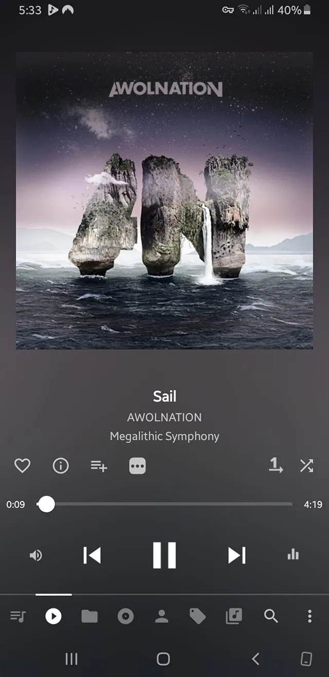 The image displays a music player interface featuring the song "Sail" by AWOLNATION from the album "Megalithic Symphony." The background of the album cover showcases a surreal landscape with large, rocky formations rising from a turbulent ocean. These formations appear to have waterfalls cascading down their sides, creating a striking visual effect. The sky is a gradient of colors, transitioning from dark to light, with stars visible, suggesting a twilight or dawn setting.

At the top of the image, the band name "AWOLNATION" is prominently displayed in bold, white letters. Below this, the song title "Sail" is listed, followed by the artist's name "AWOLNATION" again, and the album title "Megalithic Symphony." The text is clear and legible against the background.

The music player interface shows the current time at 5:33 and indicates that the song has been playing for 9 seconds. There are various icons visible, including a heart icon for favorites, a play/pause button, and a progress bar indicating the song's duration of 4 minutes and 19 seconds. The battery level of the device is at 40%, and there is a signal strength indicator in the top corner.

Overall, the image combines elements of music, art, and technology, presenting a visually engaging album cover alongside a functional music player interface.