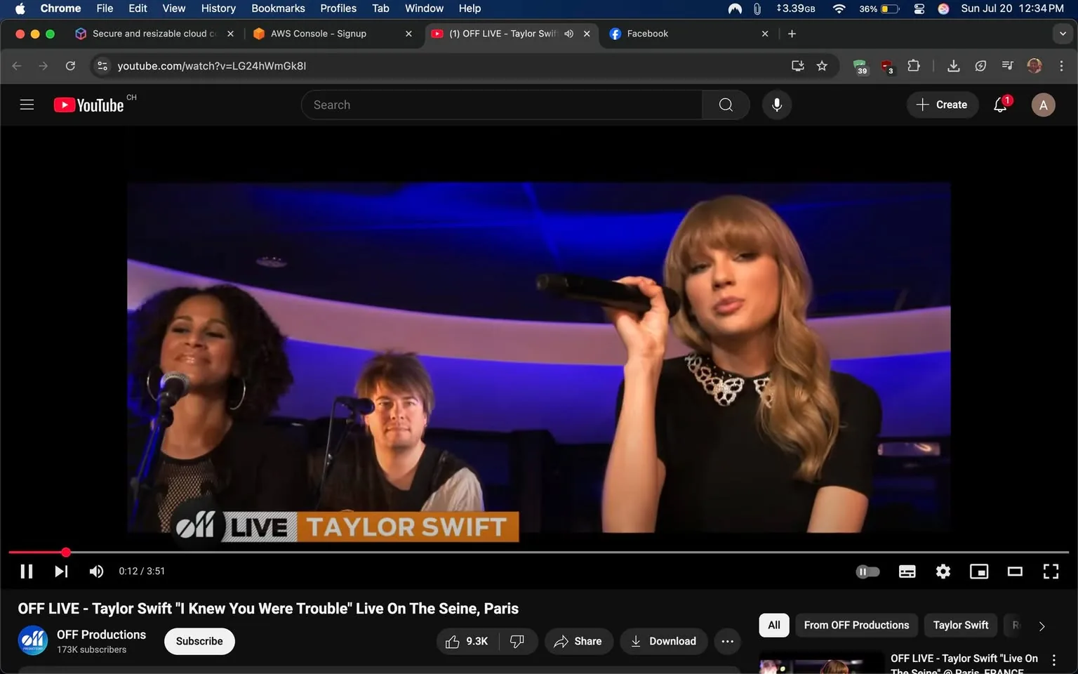 The image captures a live performance featuring three individuals on stage. The prominent figure on the right is a woman with long, wavy blonde hair, wearing a black dress adorned with a decorative collar. She holds a microphone, singing with an expressive face that conveys emotion, possibly reflecting the song's themes. 

To her left, a woman with curly hair and large hoop earrings stands at a microphone, smiling, suggesting enjoyment or engagement with the performance. In the background, a man with short, tousled hair and a casual outfit appears to be playing an instrument, likely contributing to the musical arrangement.

The setting has a modern aesthetic, with a softly lit background featuring blue and purple hues, creating an intimate atmosphere. The text at the bottom indicates this is a live performance of "I Knew You Were Trouble" by Taylor Swift, filmed on the Seine in Paris, suggesting a romantic or nostalgic context. The overall mood is lively and engaging, with the performers connecting with the audience through their expressions and gestures.