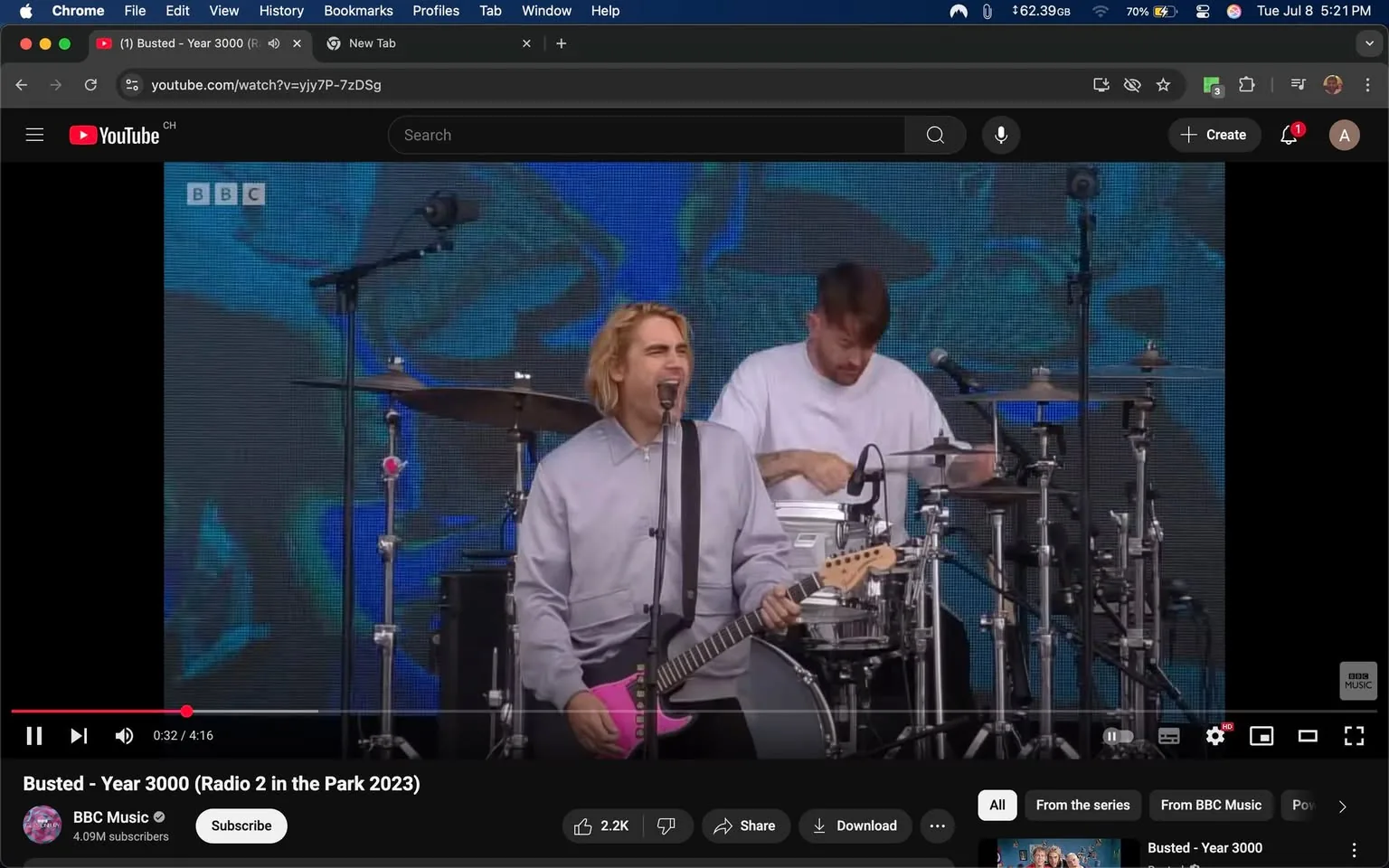 In the image, a male musician stands at the forefront, passionately singing into a microphone. He has shoulder-length, light blonde hair and is wearing a loose, gray shirt. His guitar is notably pink, contrasting with his outfit. Behind him, a drummer is visible, focused on playing, wearing a plain white shirt. The backdrop features vibrant blue and green patterns, adding energy to the scene. The overall mood appears lively and engaging, typical of a live performance. The positioning of the singer in front of the drummer suggests a dynamic interaction between the two, emphasizing the collaborative nature of the performance.