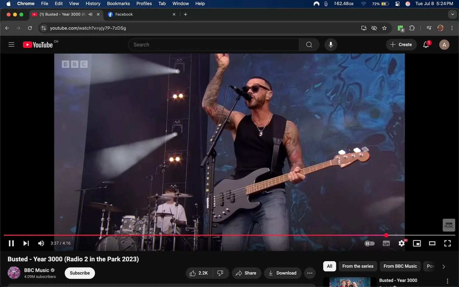 In the image, a male musician stands prominently on stage, holding a bass guitar. He has a muscular build and is wearing a sleeveless black shirt that accentuates his tattoos, which cover both arms. The tattoos are intricate and varied, suggesting a personal story or artistic expression. He wears dark sunglasses, which add a cool, confident vibe to his appearance. His expression is animated, with his mouth open as if singing or engaging with the audience, conveying a sense of energy and enthusiasm.

The musician is positioned slightly to the left of the frame, with his right arm raised, possibly gesturing to the crowd or emphasizing a point in the performance. His left hand grips the neck of the bass guitar, which is a sleek black instrument with visible strings and a wooden neck. The stage is illuminated by bright lights, with beams of light cutting through the air, creating a dynamic atmosphere. In the background, there is a large screen displaying abstract visuals, enhancing the visual experience of the performance.

Behind him, a drummer is partially visible, dressed in a white shirt, contributing to the band's sound. The drummer appears focused, with a drum kit that includes a snare, cymbals, and a bass drum, indicating a full band setup. The overall scene suggests a live concert environment, likely filled with fans, as the energy from the musician implies an engaging performance.

The lighting is bright and vibrant, contributing to a festive and lively mood. The combination of the musician's confident posture, the dynamic lighting, and the abstract visuals creates an atmosphere of excitement and celebration. The image captures a moment of performance that is likely part of a larger event, possibly a music festival, given the context of the title "Radio 2 in the Park 2023."

There are no visible logos or branding on the musician's attire, but the presence of the BBC logo in the upper left corner indicates that this performance is associated with the BBC Music platform. The overall tone of the image is energetic and celebratory, reflecting the joy of live music and the connection between the performer and the audience.