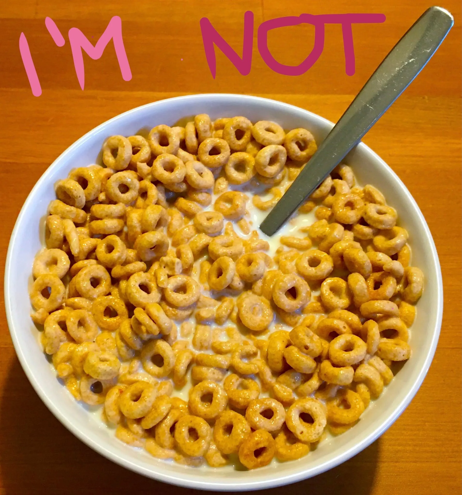 The image features a bowl of cereal, specifically a type of O-shaped breakfast cereal, submerged in milk. The bowl is white and is placed on a wooden table, which has a warm, natural finish. A silver spoon rests in the bowl, partially submerged among the cereal pieces. 

Above the bowl, there is text that reads "I'M NOT" in a playful, pink font, suggesting a humorous or lighthearted tone. The text's placement indicates that it might be a response to a common question or assumption, possibly about the cereal itself or breakfast choices.

There are no identifiable people, actions, or gestures in the image. The focus is entirely on the cereal and the text. The overall mood seems casual and whimsical, likely intended to evoke a smile or chuckle from viewers. There are no explicit, controversial, or sensitive content elements present in the image.