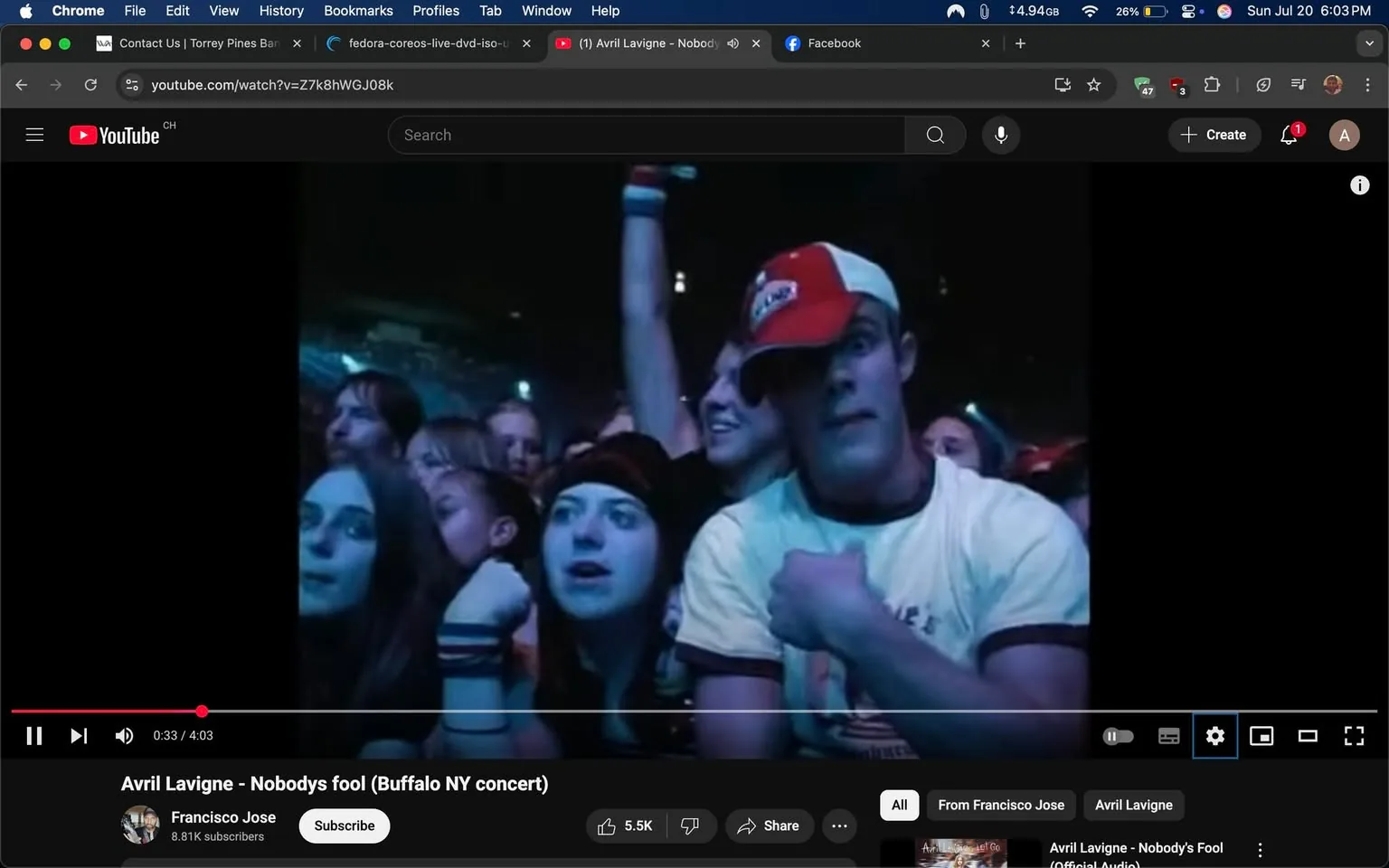 The image captures a vibrant moment from a concert, likely featuring Avril Lavigne, as indicated by the title in the video player. The scene is filled with energy and excitement, characteristic of live music events. 

In the foreground, a group of fans is visible, their expressions reflecting a mix of enthusiasm and anticipation. The lighting is dim, with blue hues illuminating the crowd, creating a lively yet intimate atmosphere. 

On the left side, a young woman with long, dark hair and a black top appears to be singing along, her face showing a blend of concentration and joy. Next to her, another fan, wearing a black beanie, looks equally engaged, her mouth slightly open as if caught in the moment of the performance. 

To the right, a young man stands out, wearing a white shirt with a red and white cap. His expression is intense, possibly caught up in the music or the excitement of the crowd. He seems to be flexing his arm, which adds to the energetic vibe of the scene. His posture suggests he is fully immersed in the experience, embodying the thrill of being at a live concert. 

Behind them, other concert-goers can be seen, some with their arms raised, contributing to the collective energy of the crowd. The background features a mix of faces, all focused on the stage, showcasing the communal experience of live music. 

The overall tone of the image is one of exhilaration and connection, as fans come together to enjoy the performance. The visual elements—expressive faces, dynamic poses, and the atmospheric lighting—convey a sense of shared joy and passion for music. 

This moment encapsulates the essence of a concert: the thrill of live performance, the connection between the artist and the audience, and the unforgettable memories created in such vibrant settings.